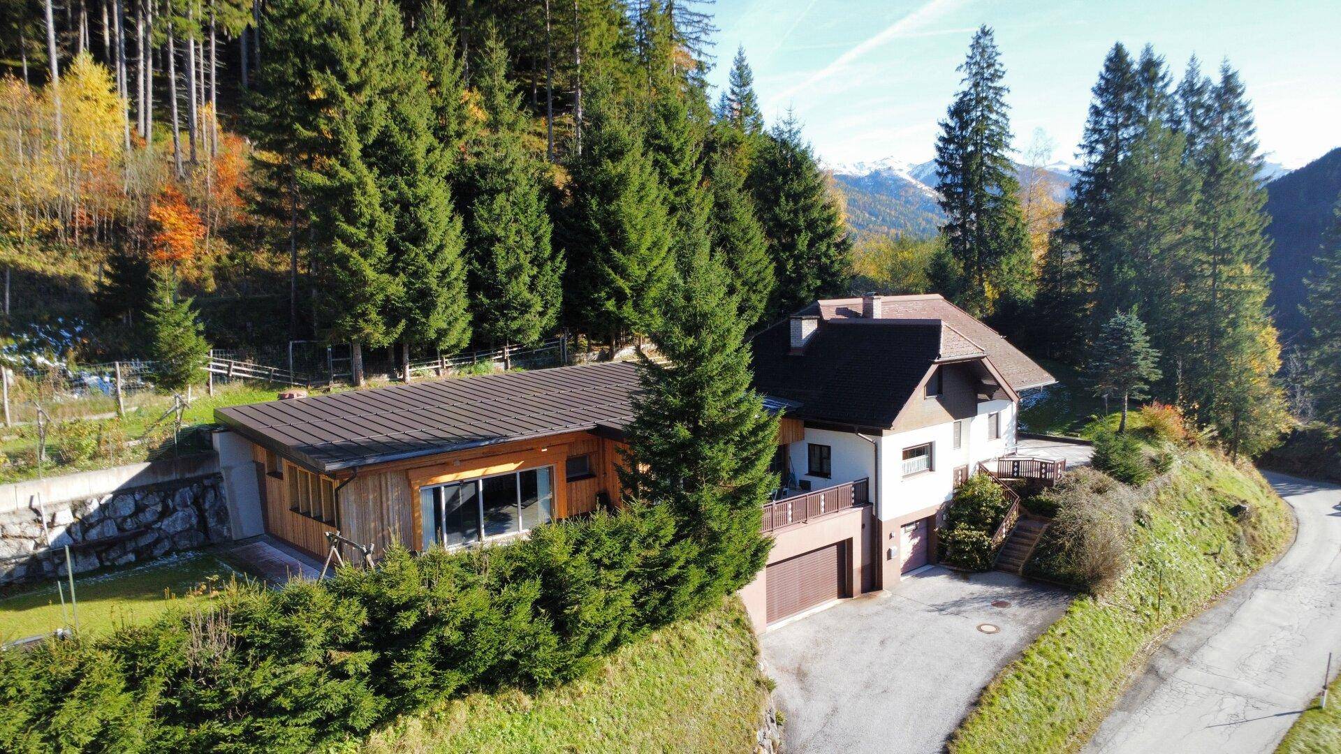 Luftaufnahme eines Hauses in Hanglage mit Anbau und umgebender Natur und Bergen.