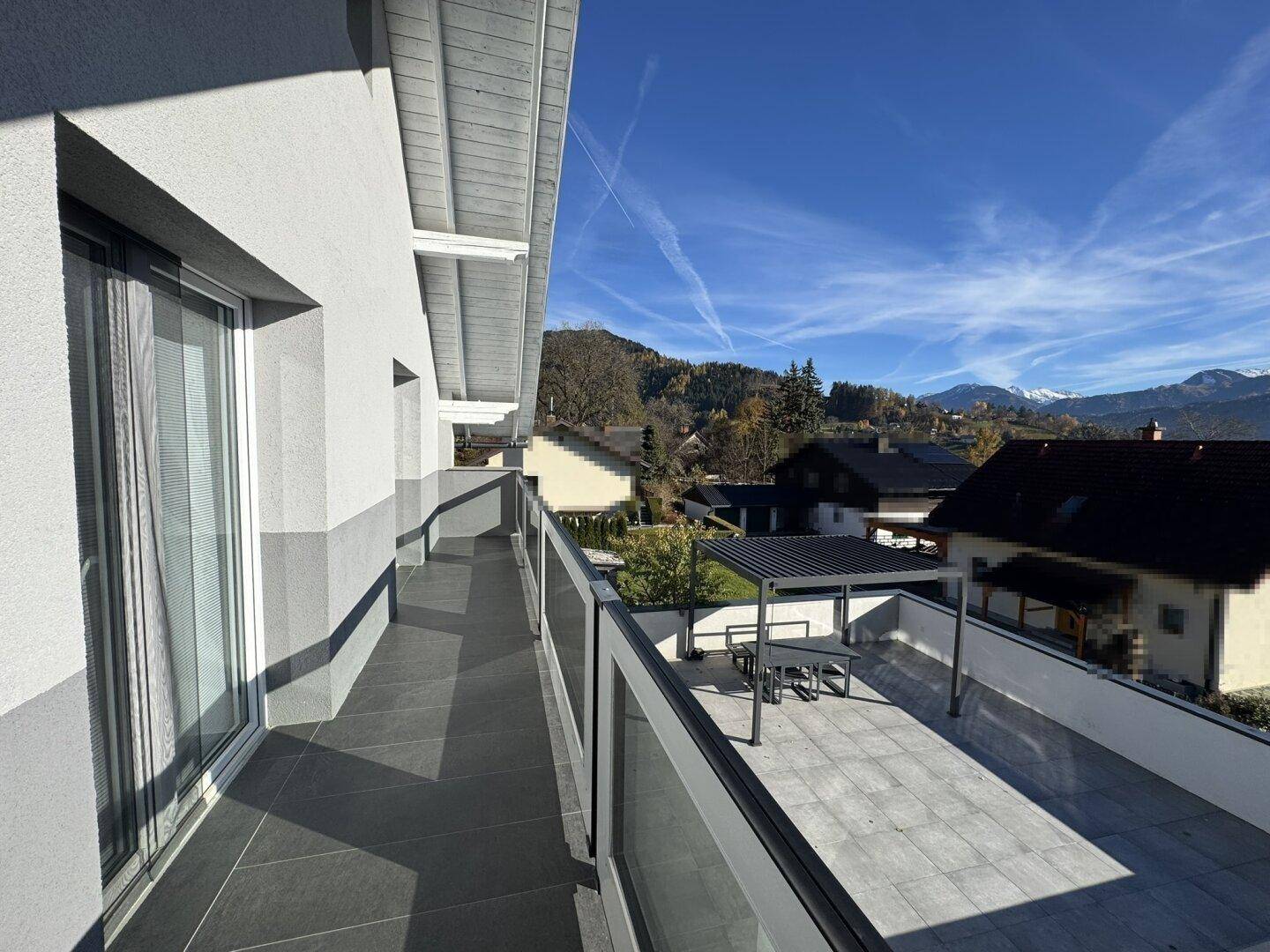 Langer Balkon mit modernem Glasgeländer, bietet Zugang zur Terrasse und Bergblick.