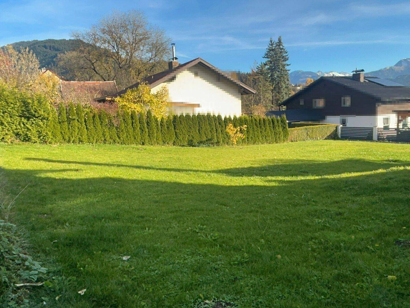 Sonnige Rasenfläche mit gepflegter Hecke, umgeben von Häusern und einem weiten Bergblick.