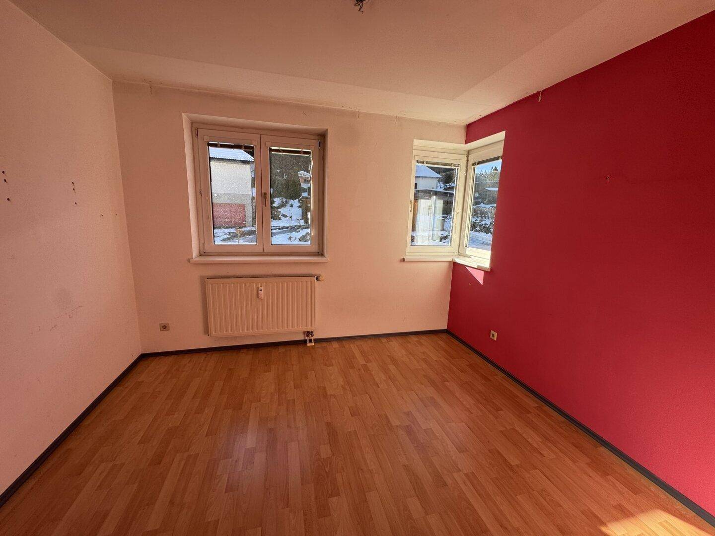 Leeres Zimmer mit Holzboden, zwei Fenstern und einer auffälligen roten Akzentwand.