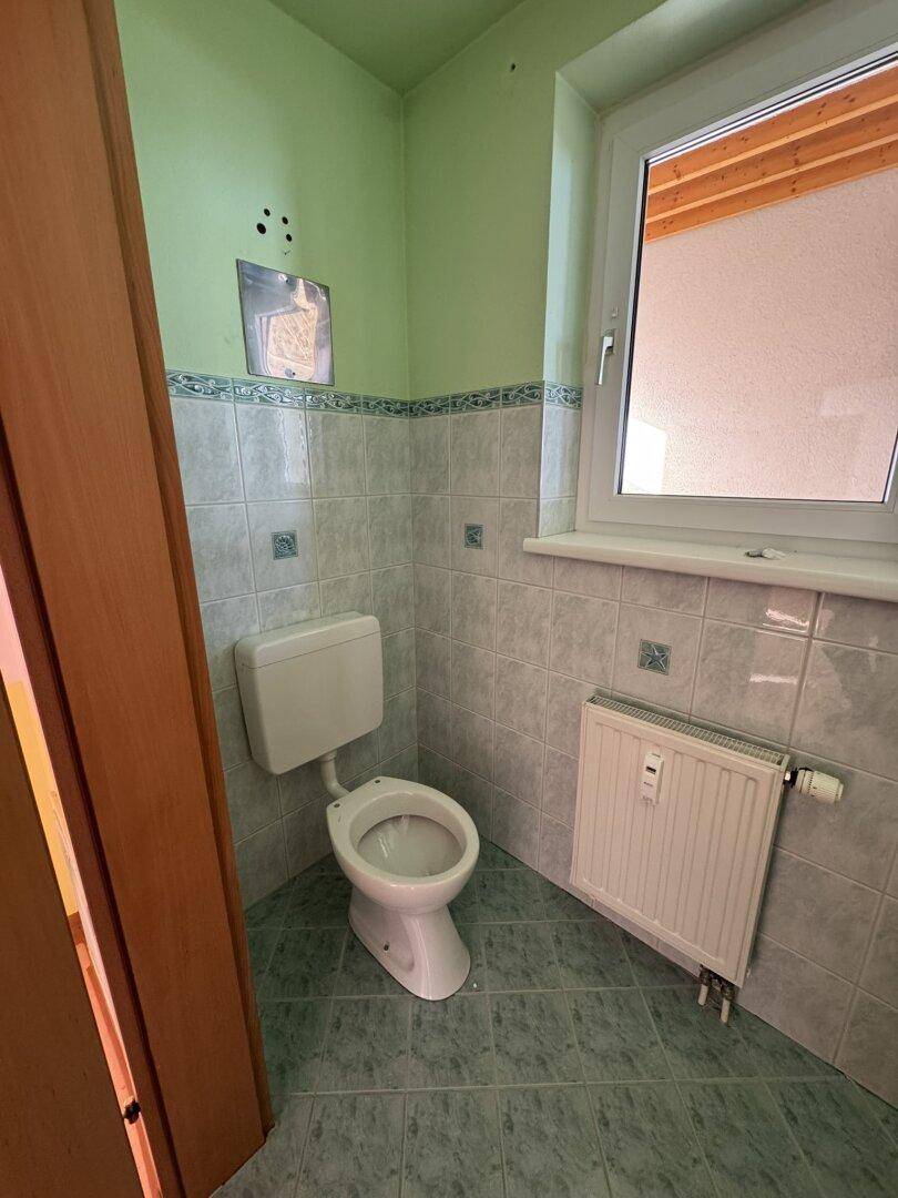 Kleines WC mit grünen Fliesen, Toilette und Fenster, das eine Renovierung benötigt.