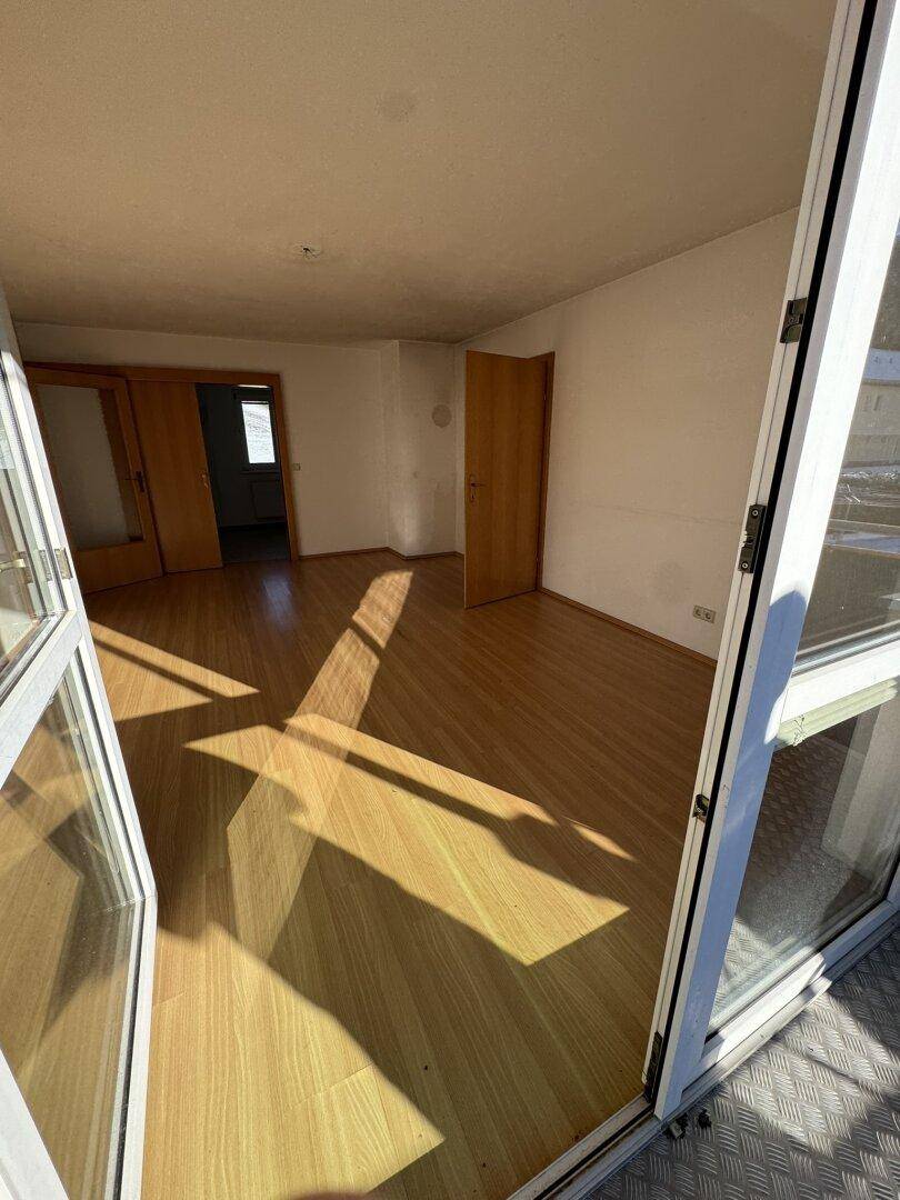 Geräumiges Zimmer mit Holzboden und offener Balkontür, die viel Sonnenlicht hereinlässt.