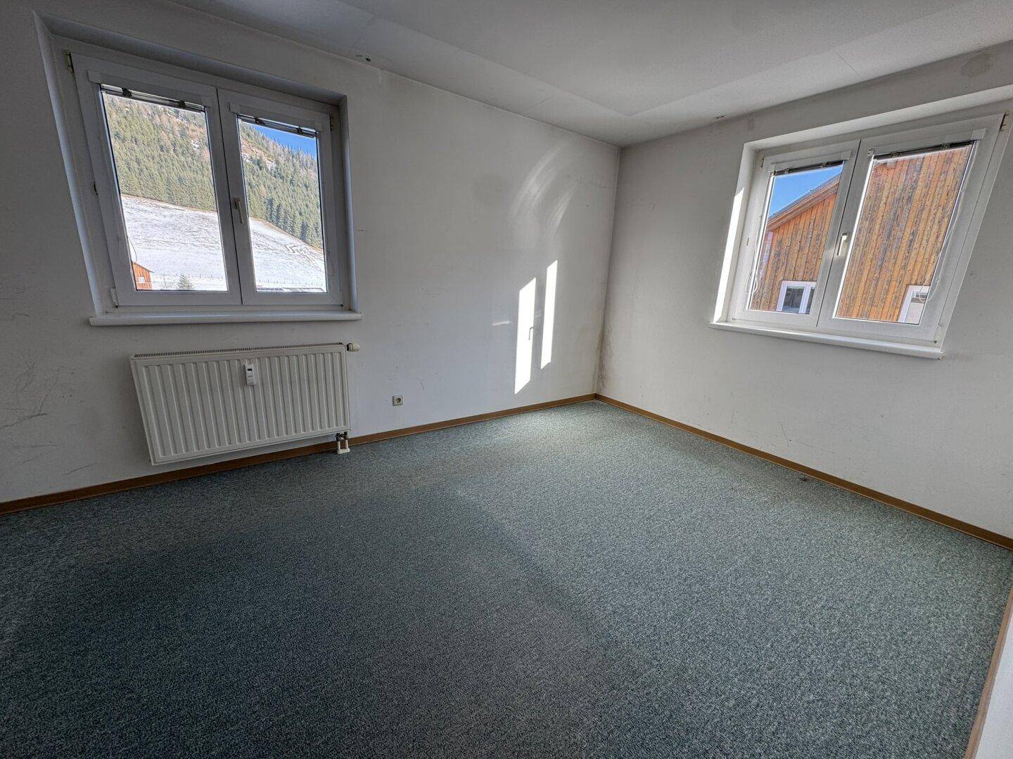 Leeres Zimmer mit blauem Teppichboden und Fenstern mit Blick auf die verschneiten Berge.