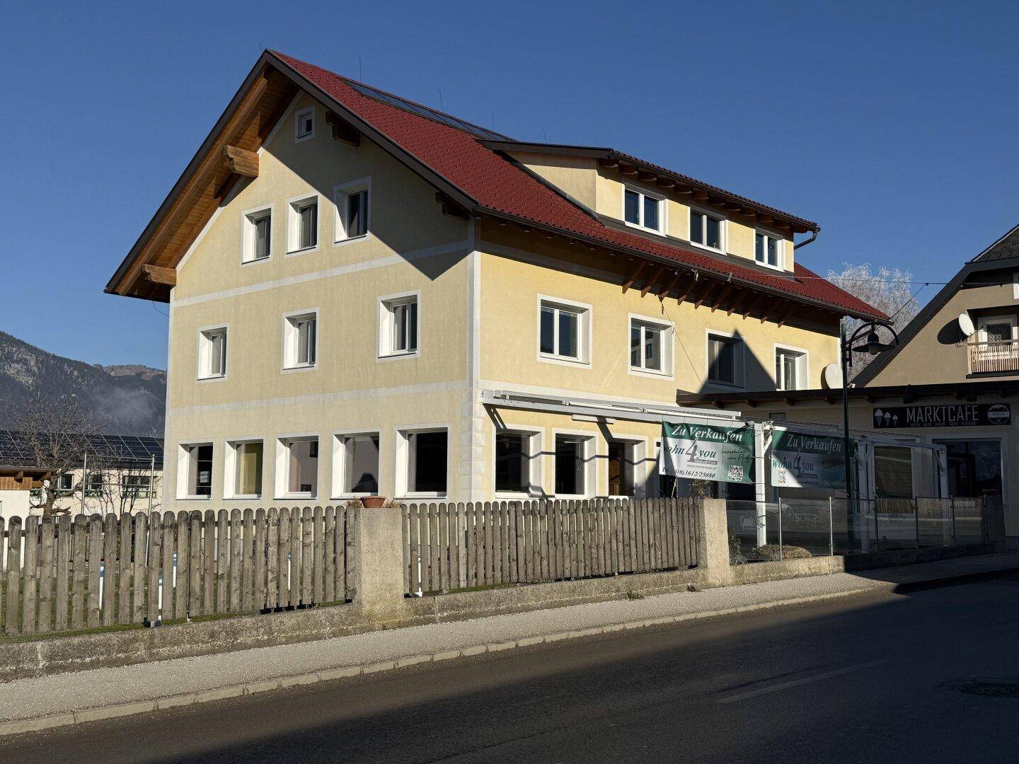 Vorderansicht des gelben Gebäudes mit rotem Dach, Holzzaun und Photovoltaikanlage auf dem Dach.