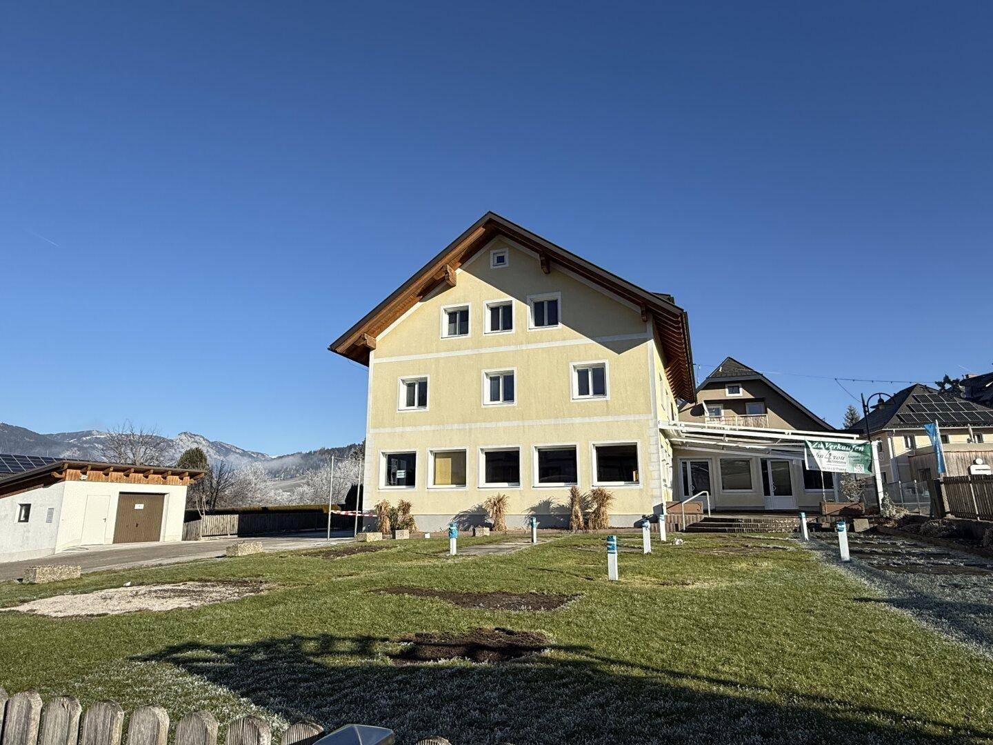 Gelbes Gebäude mit Satteldach und großem Grundstück im Winter, umgeben von Bergen unter blauem Himmel.