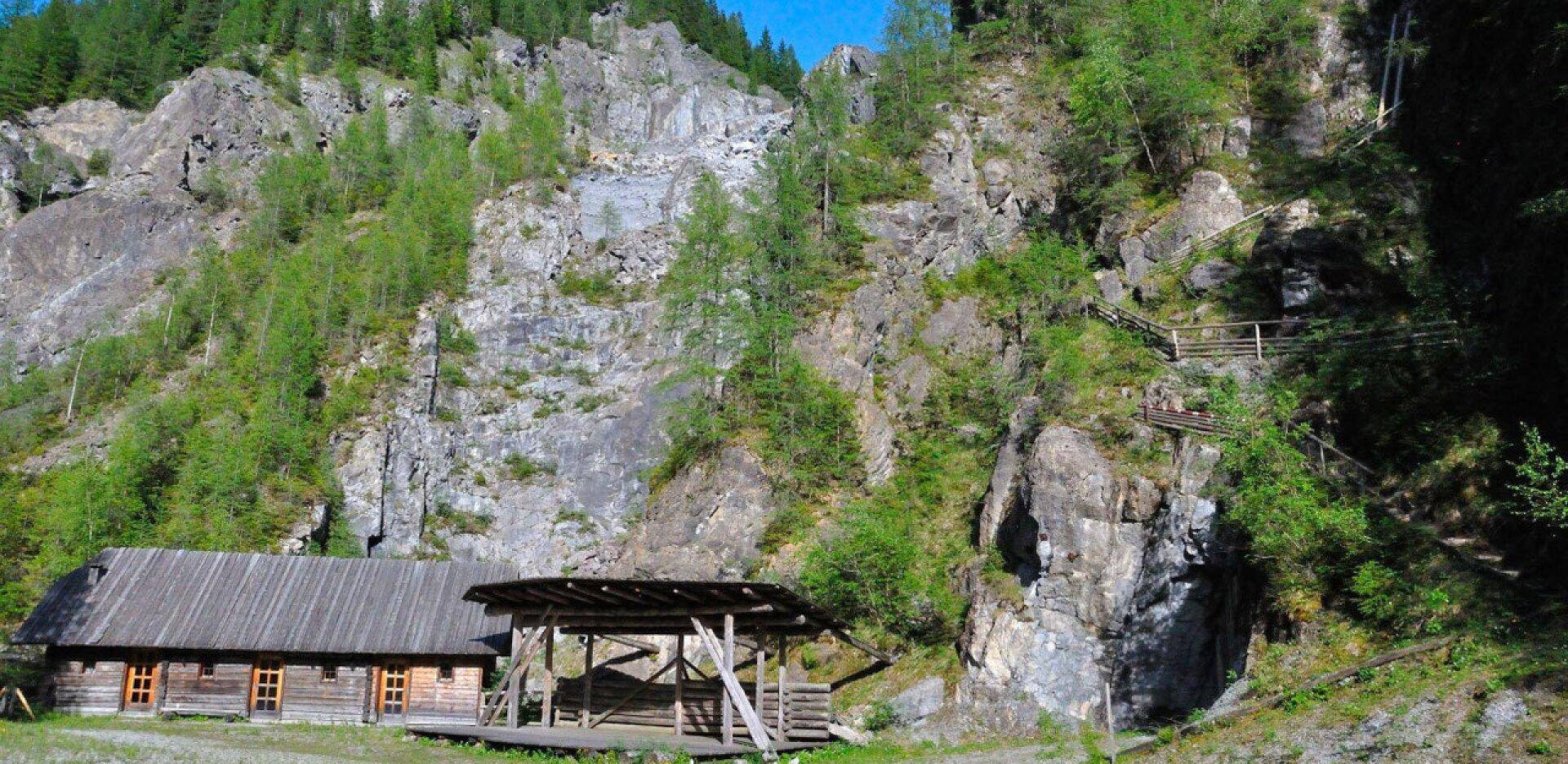Rustikales Holzgebäude am Fuße einer steilen Felswand, umgeben von Bäumen und Wanderwegen.