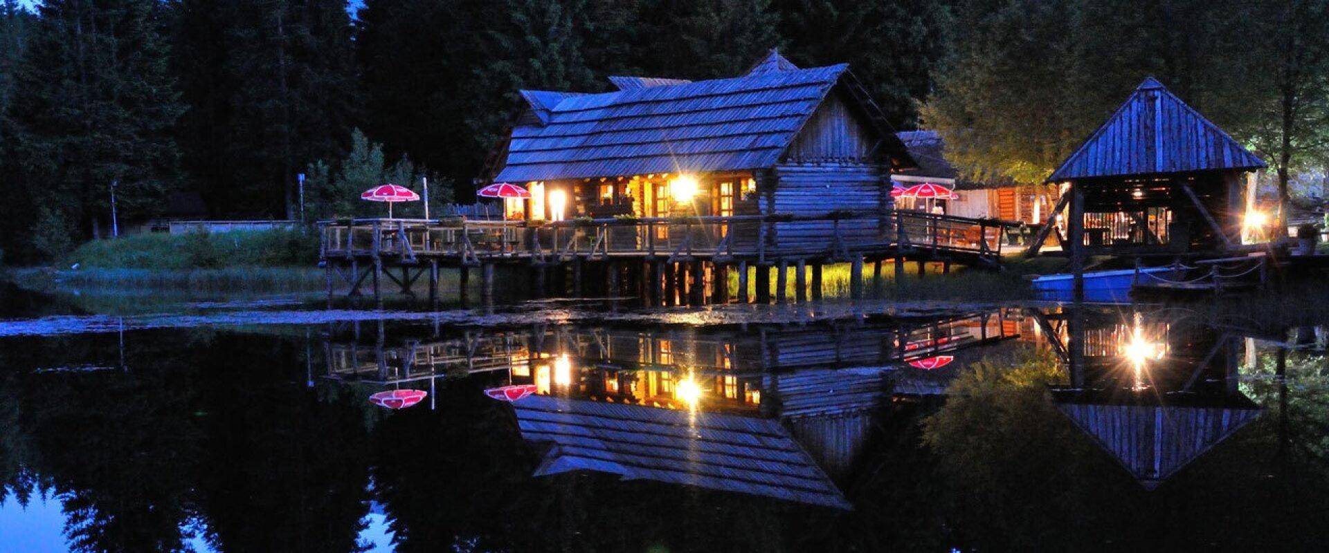 Abendliche Ansicht des beleuchteten Blockhauses am See, dessen warmes Licht sich im dunklen Wasser spiegelt.