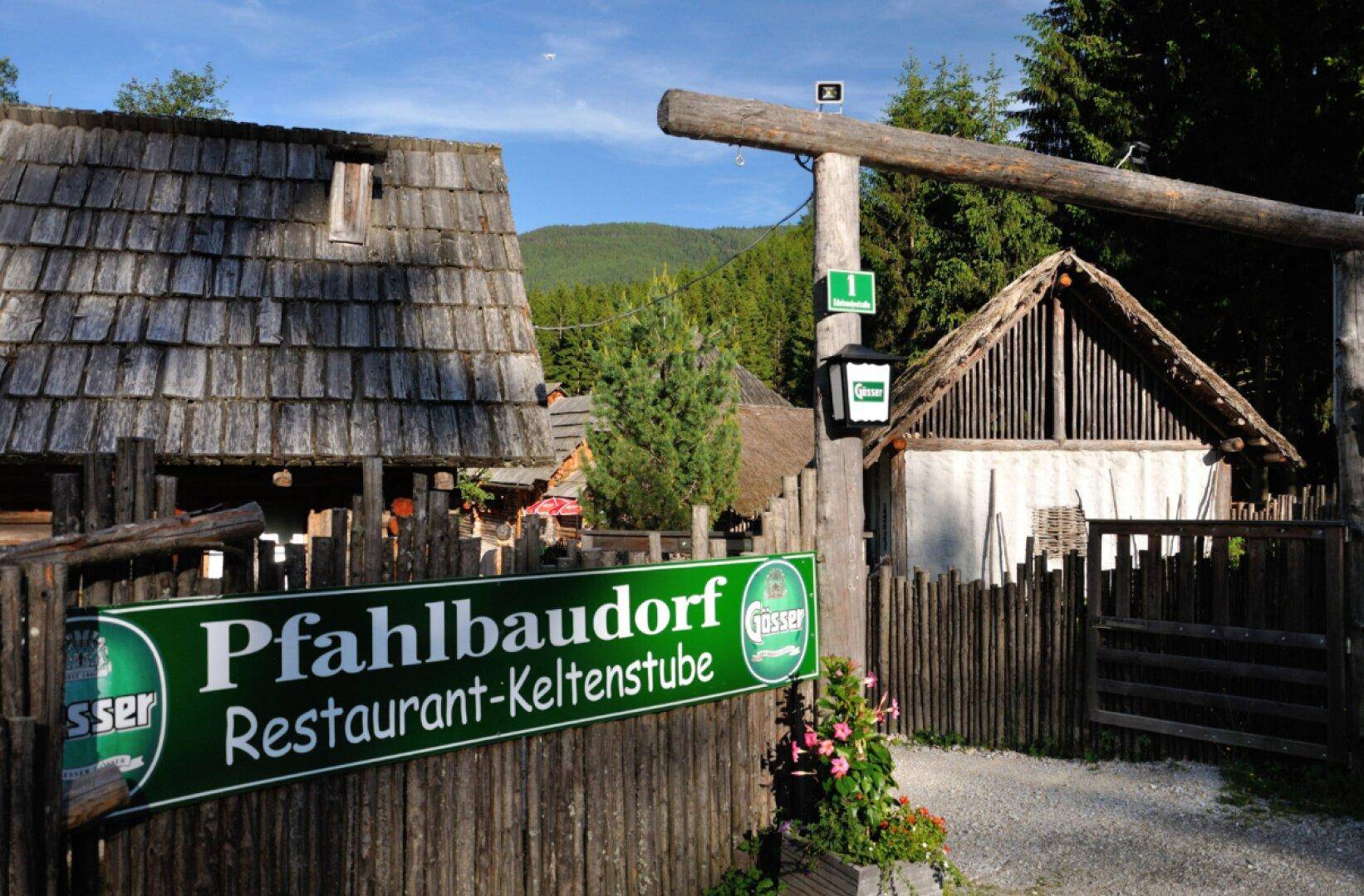 Eingangsbereich des Pfahlbaudorf Restaurants mit rustikalem Holzzaun und Beschilderung, umgeben von Bäumen.