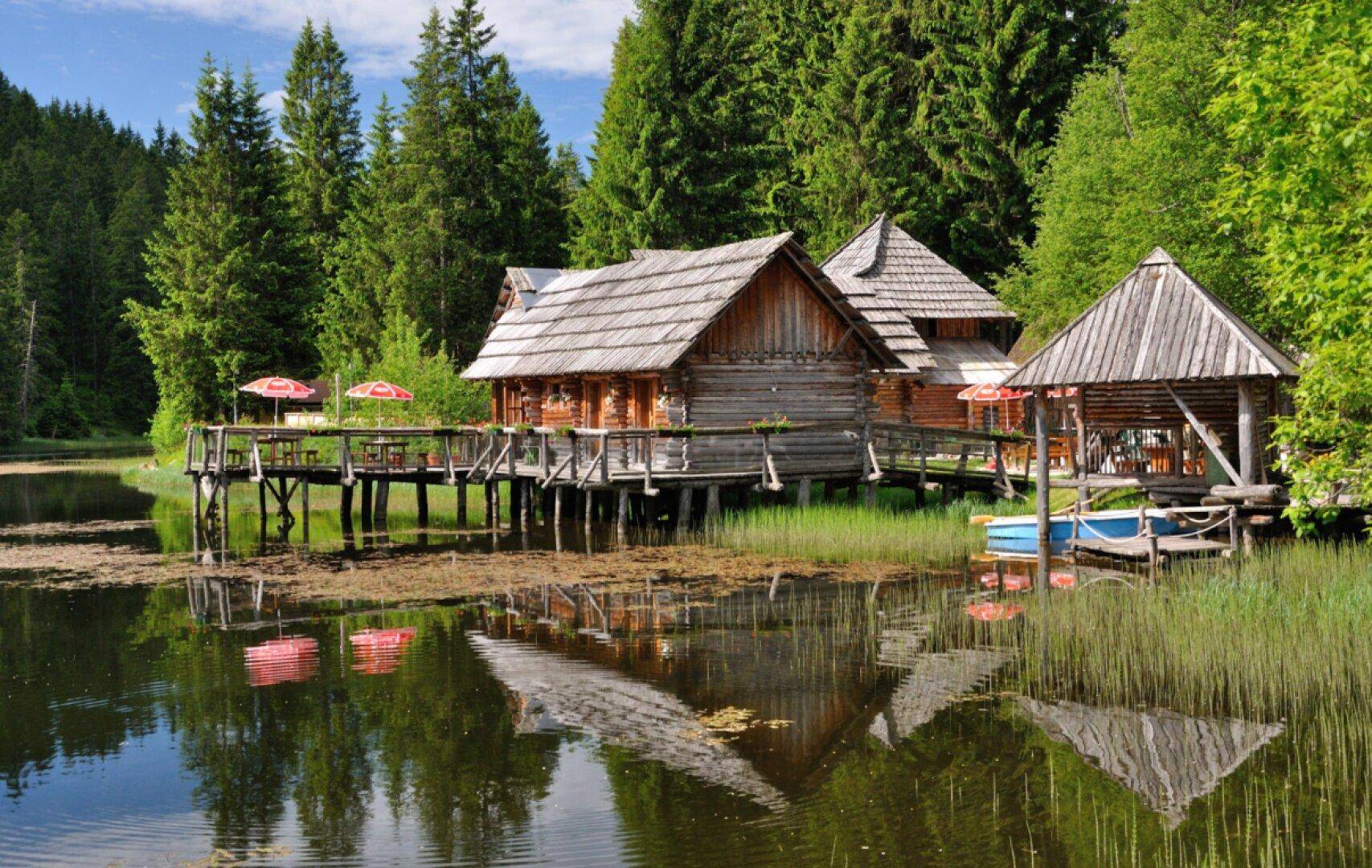 Blockhäuser am Seeufer, umgeben von üppiger Vegetation und Schilf, spiegeln sich im ruhigen Wasser.