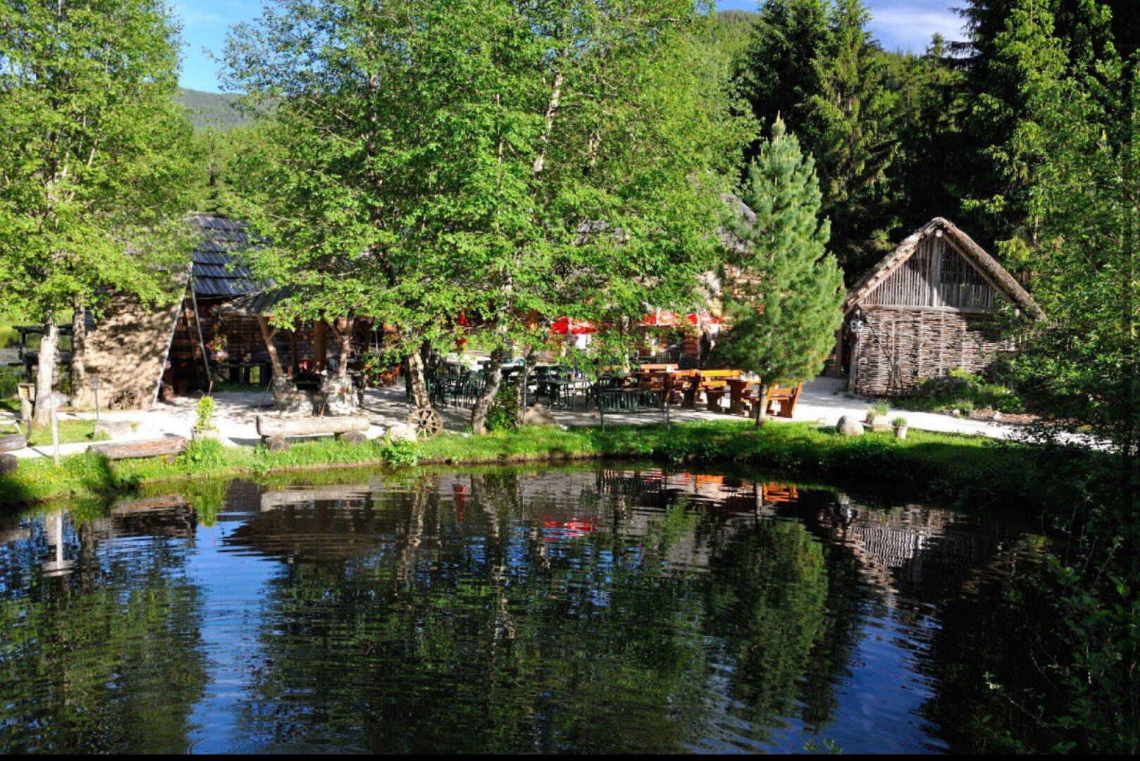 Idyllischer Außenbereich mit einem kleinen Teich, umgeben von Bäumen und Sitzgelegenheiten, im Sonnenlicht.