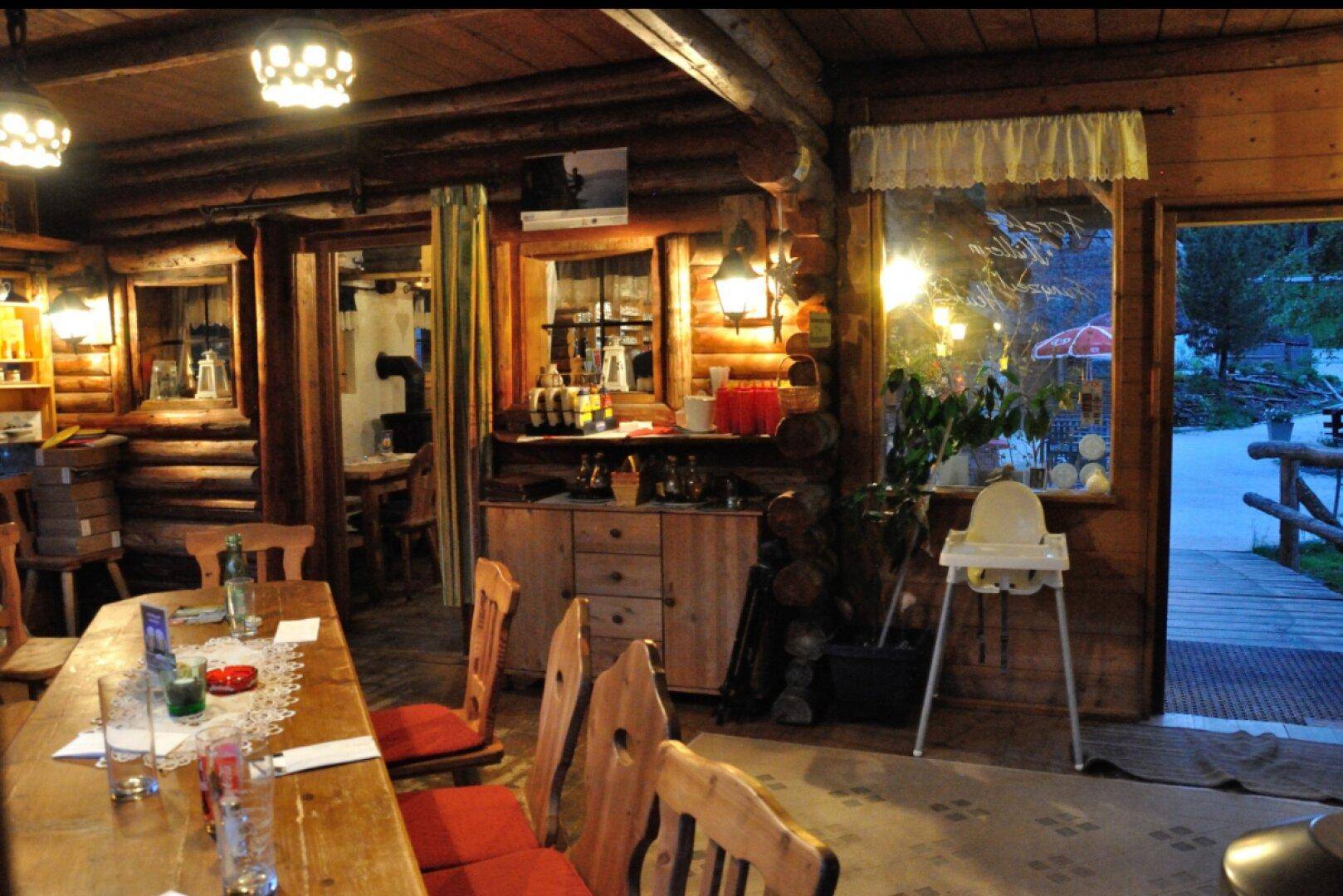 Gemütlicher Innenraum eines Blockhauses mit Holztischen, Stühlen und warmem Licht, Blick nach draußen.