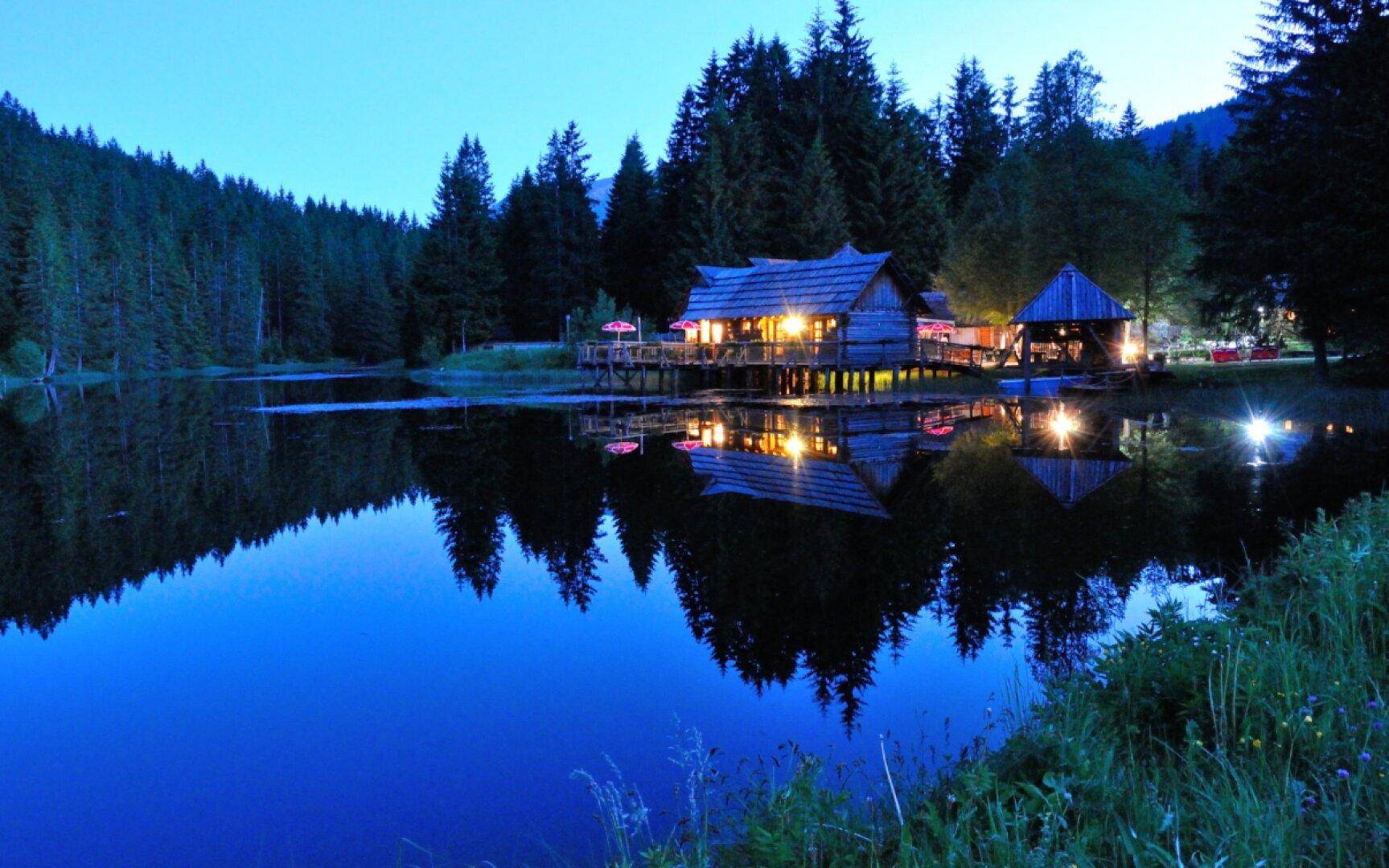 Abendstimmung am See mit beleuchtetem Blockhaus, dessen Lichter sich im dunklen Wasser spiegeln, umgeben von Wald.