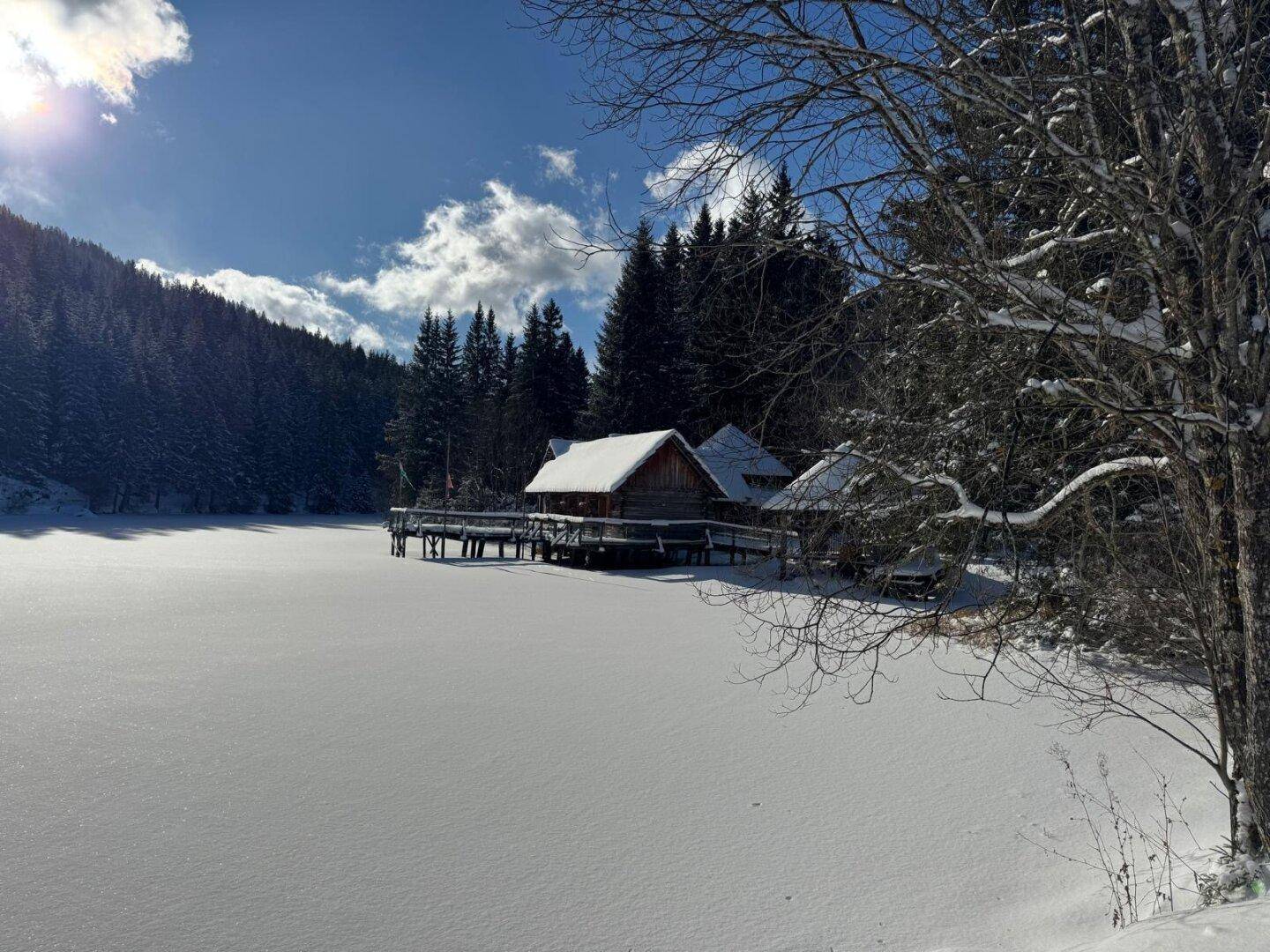 Weitläufige Winterlandschaft mit schneebedecktem See und Blockhäusern am Ufer, umgeben von verschneiten Wäldern.