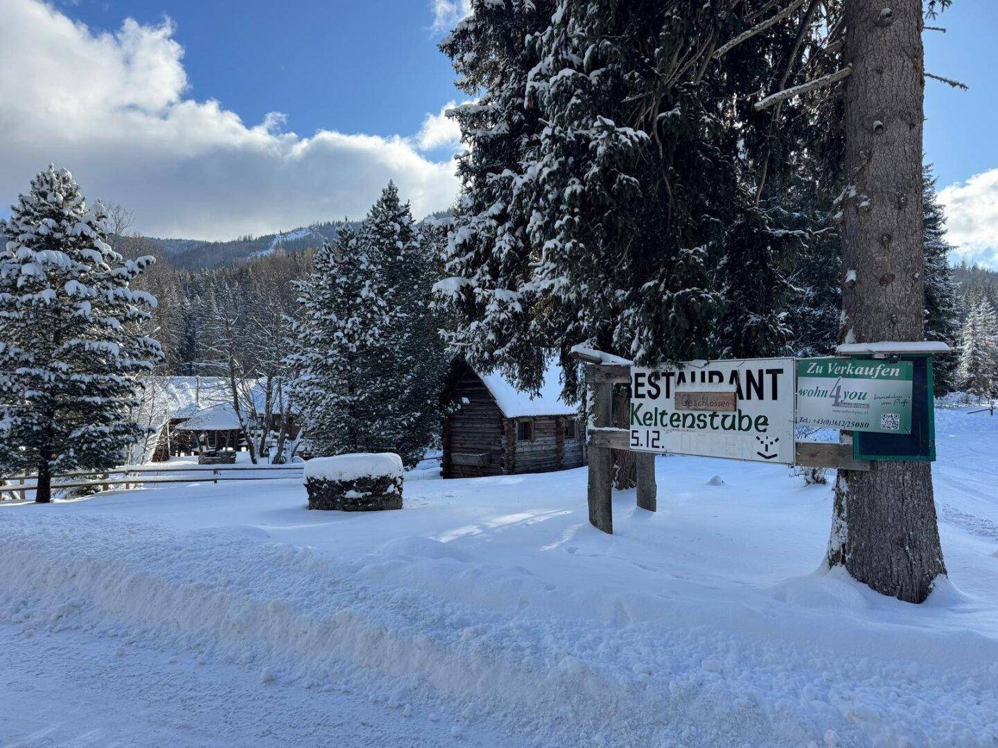 Winterlicher Eingangsbereich mit verschneiten Bäumen und einem Schild zum Restaurant, umgeben von tiefem Schnee.