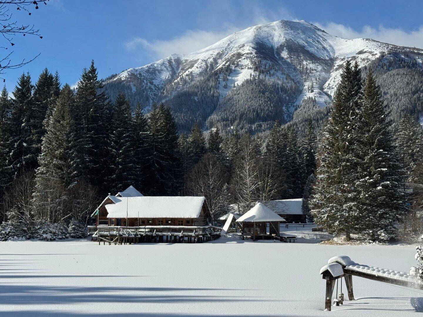 Winterliche Szenerie mit schneebedecktem Blockhaus am zugefrorenen See, umgeben von verschneiten Tannen und Bergen.