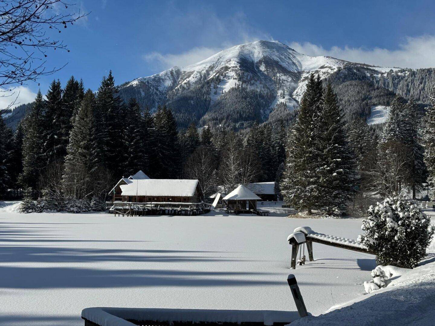 Winterlandschaft mit schneebedecktem Blockhaus am zugefrorenen See, umgeben von verschneiten Tannen und Bergen.