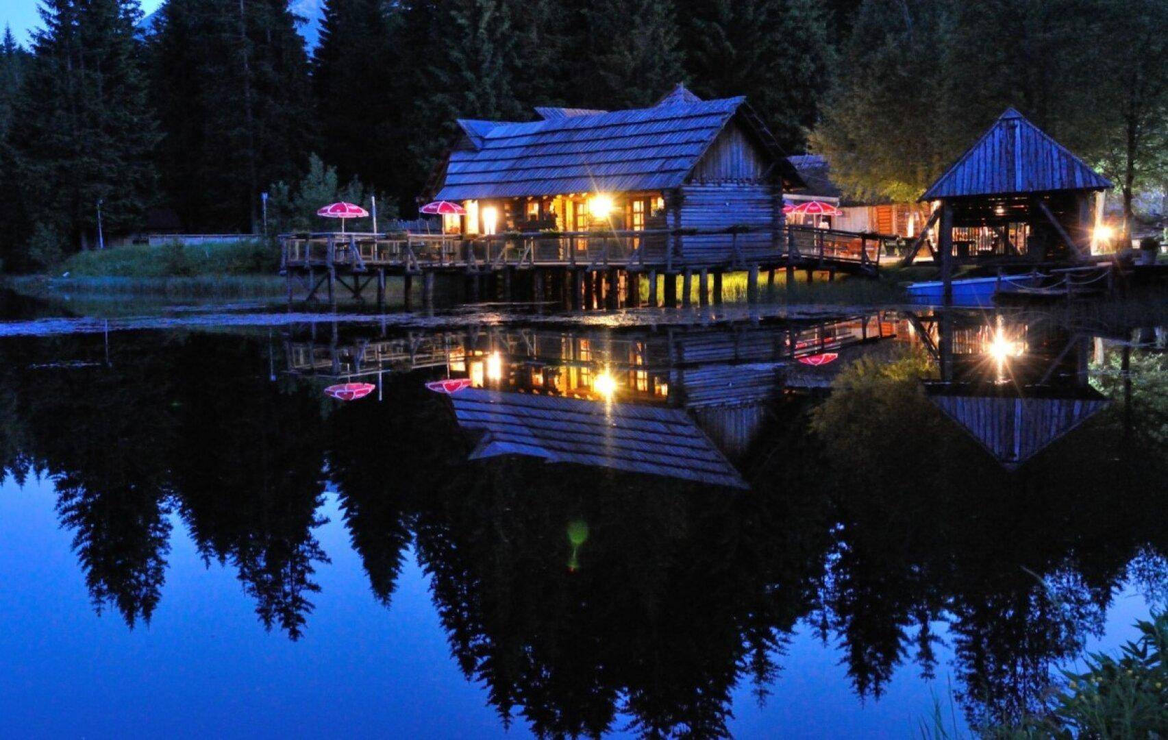 Romantische Abendstimmung am See mit beleuchtetem Blockhaus, dessen Reflexionen im Wasser tanzen.