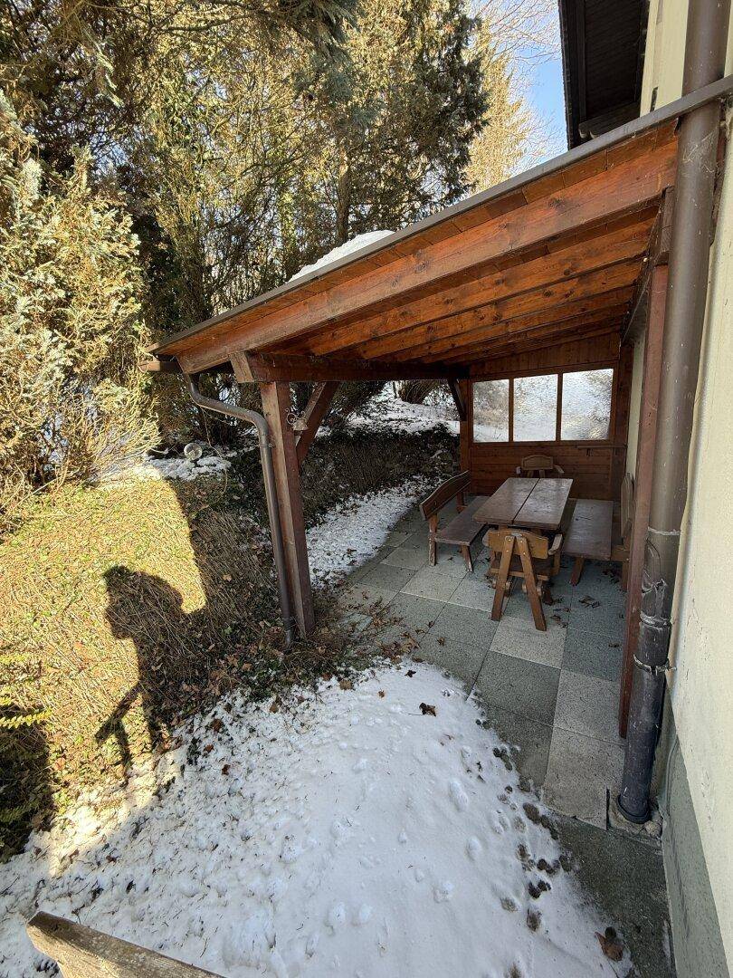 Überdachter Sitzbereich im Freien mit Holztisch und Bänken, umgeben von Schnee und Bäumen.