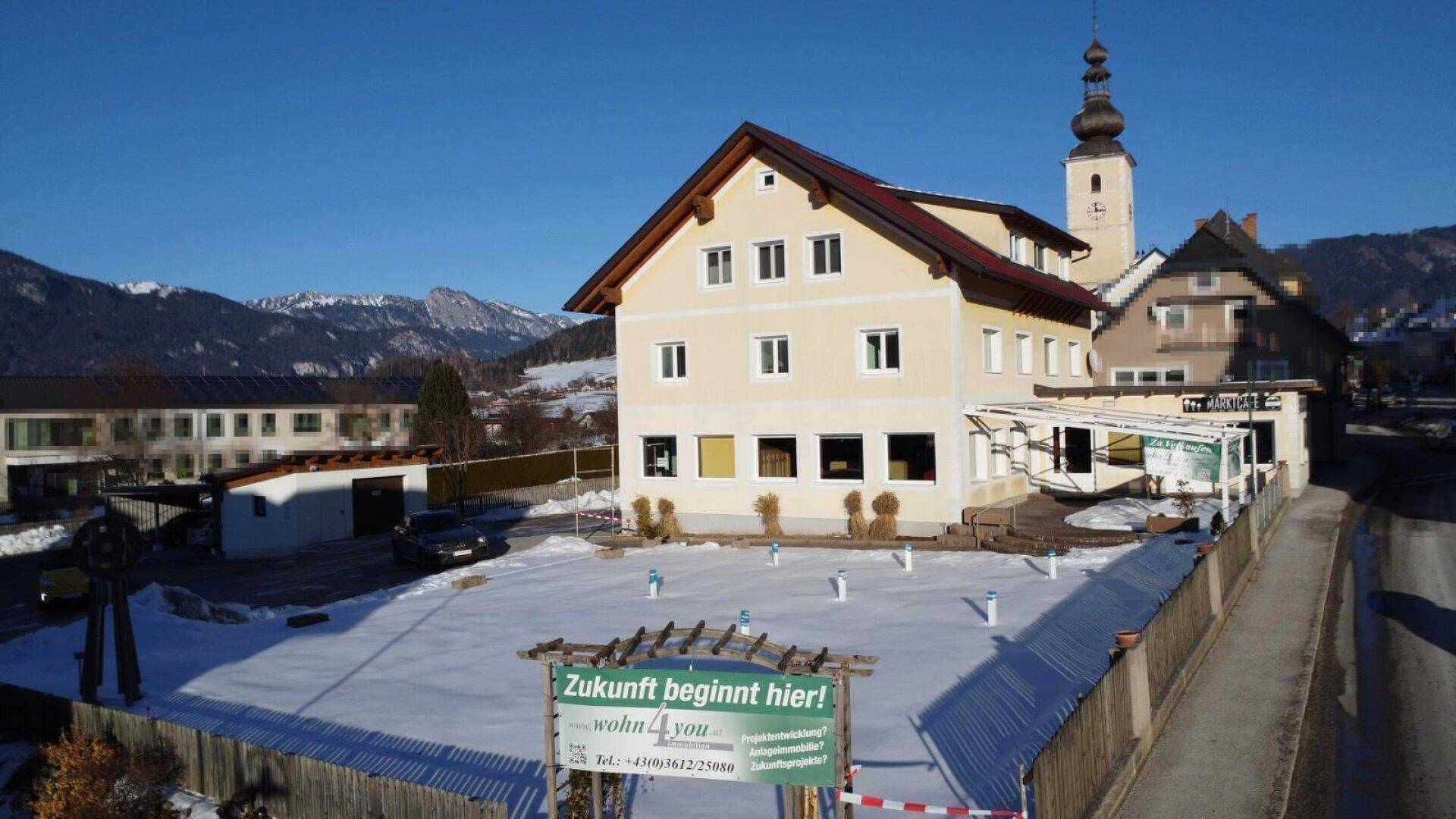 Gelbes Gebäude mit Satteldach und verschneitem Grundstück im Winter, mit Werbeschild.