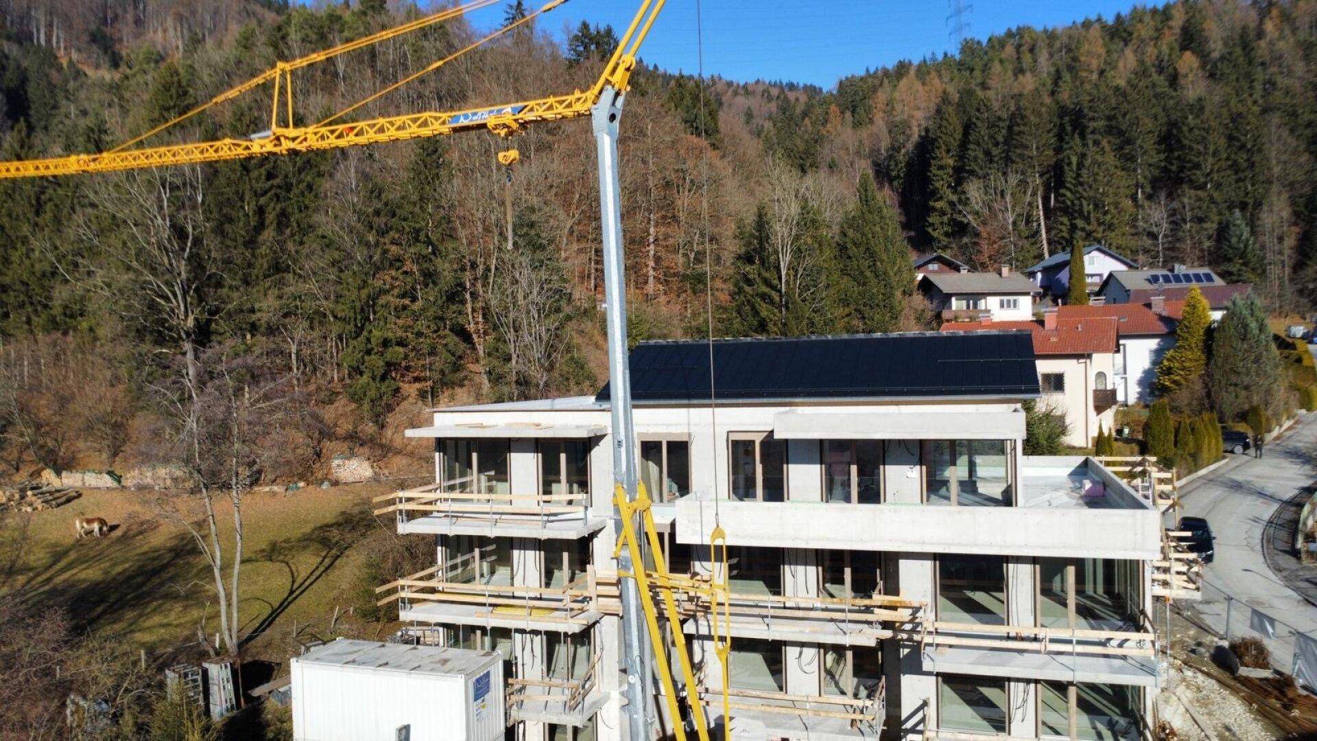 Gebäude im Bau mit Kran vor bewaldetem Hügel und angrenzender Straße.