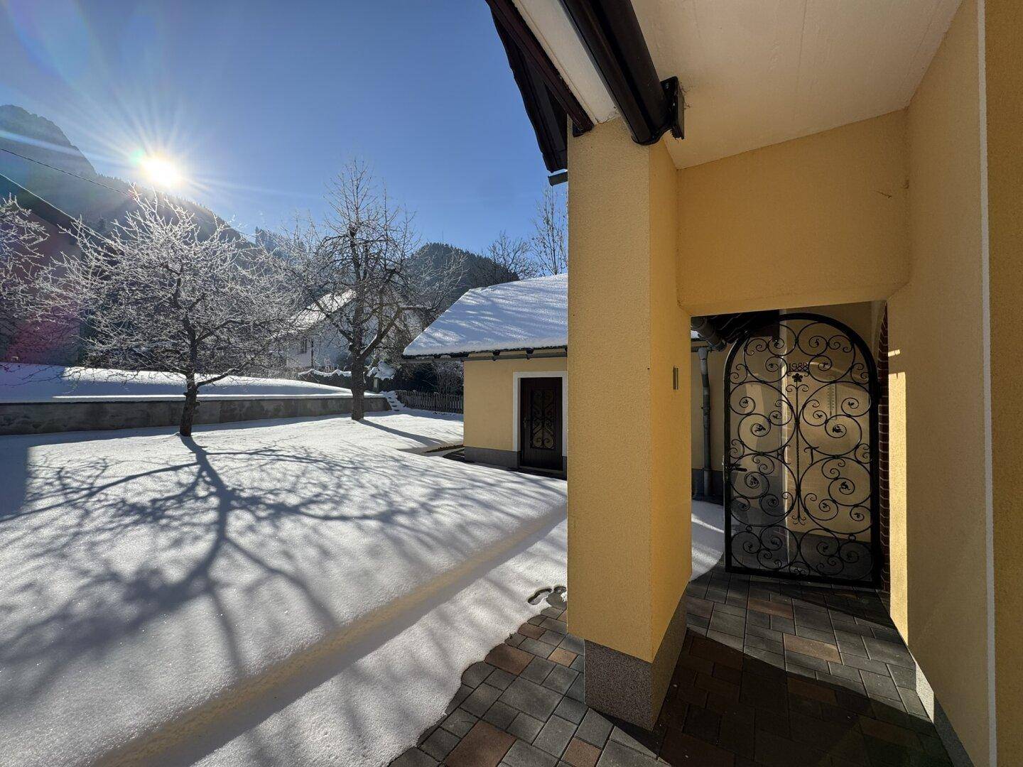 Eingangsbereich des Hauses mit gepflastertem Weg, dekorativem Tor und verschneitem Vorgarten an einem sonnigen Tag.