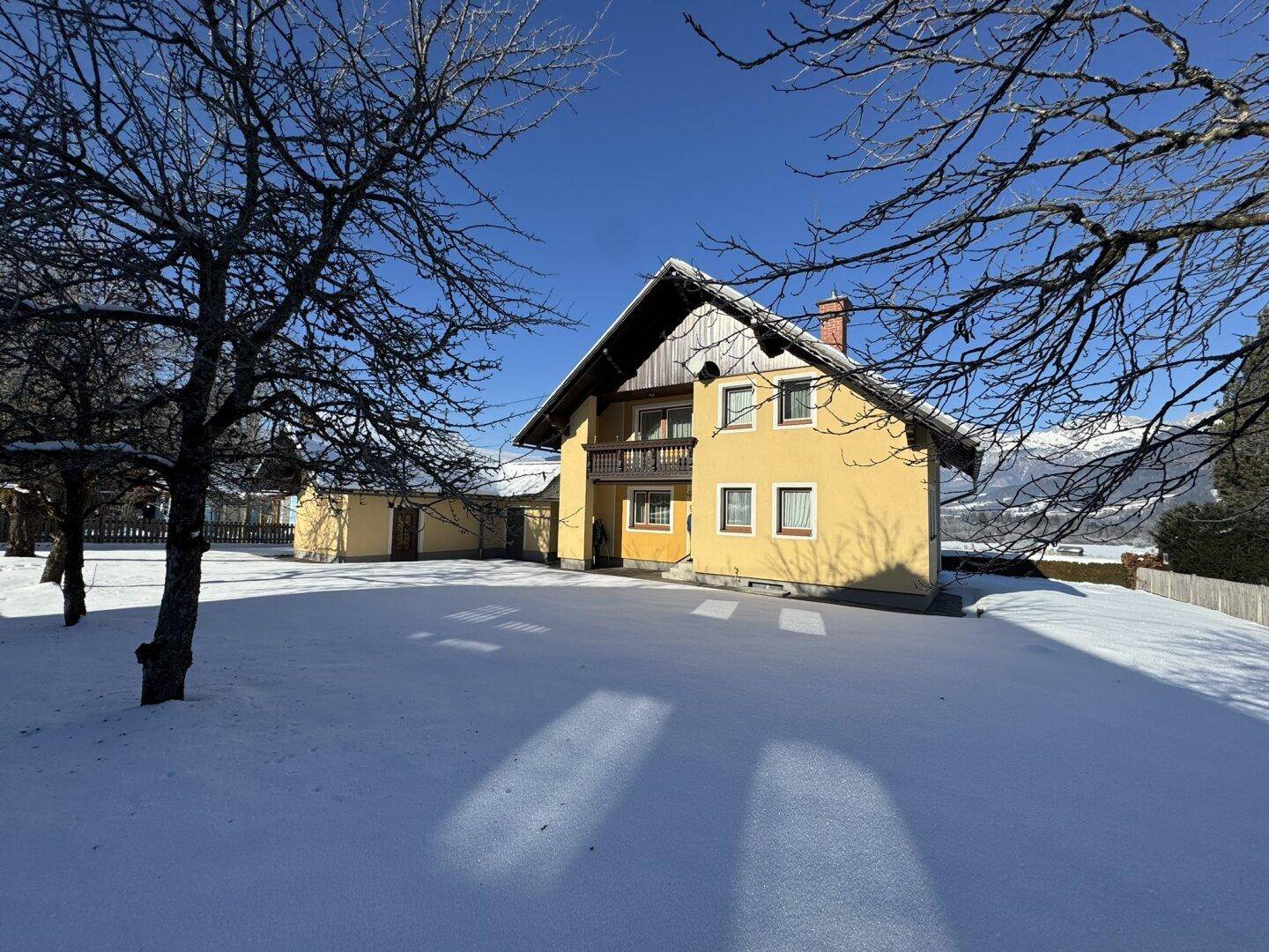 Das Haus im Winter mit verschneitem Garten und kahlen Bäumen, umgeben von einer malerischen Berglandschaft.