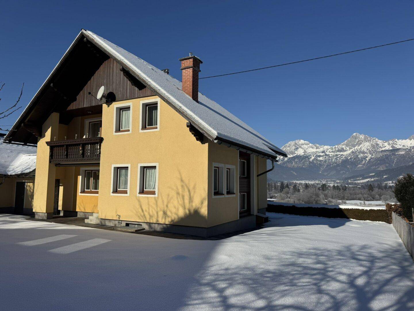 Seitenansicht des Hauses mit gelber Fassade, schneebedecktem Dach und weitem Blick auf die Berge.