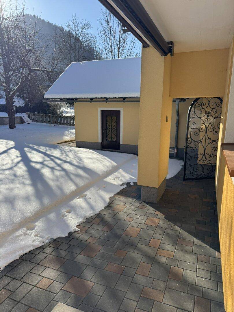 Gepflasterter Bereich neben dem Haus mit Schnee und einem kleinen Nebengebäude im winterlichen Sonnenschein.