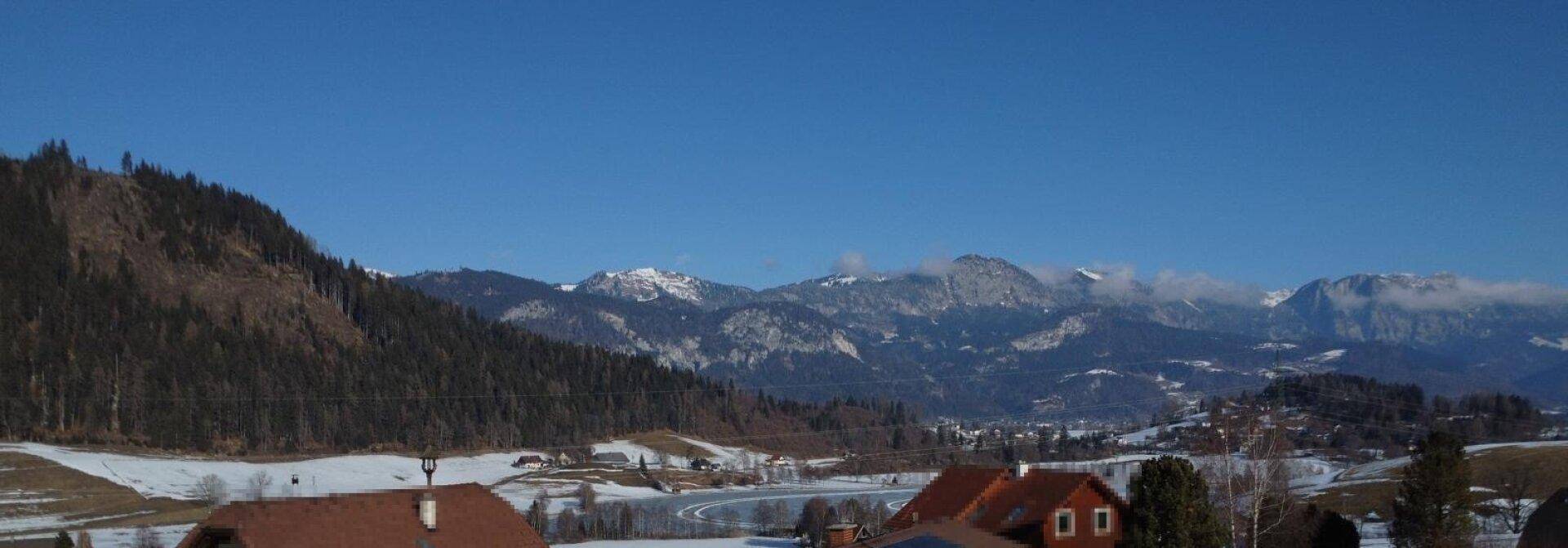 Weitläufiger Panoramablick über eine schneebedeckte Landschaft mit Bergen und vereinzelten Häusern.
