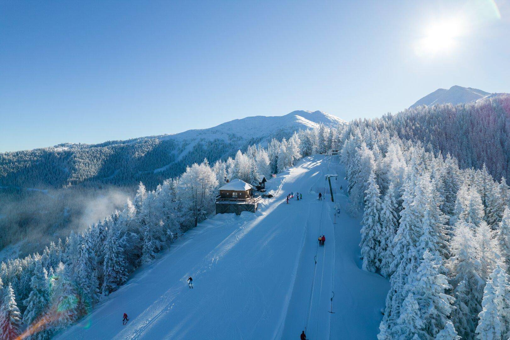 Sonnige Skipiste mit einer Berghütte und Skifahrern, umgeben von tief verschneiten Tannenbäumen.