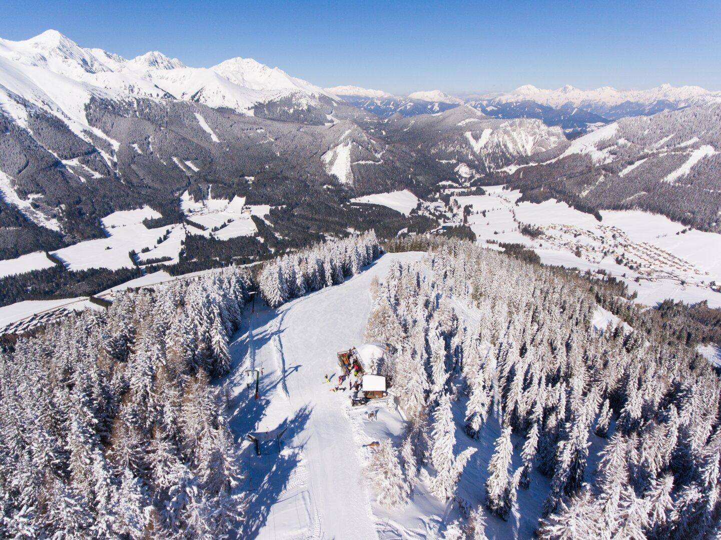 Atemberaubende Luftaufnahme einer verschneiten Berglandschaft mit dichten Wäldern und Skipisten.