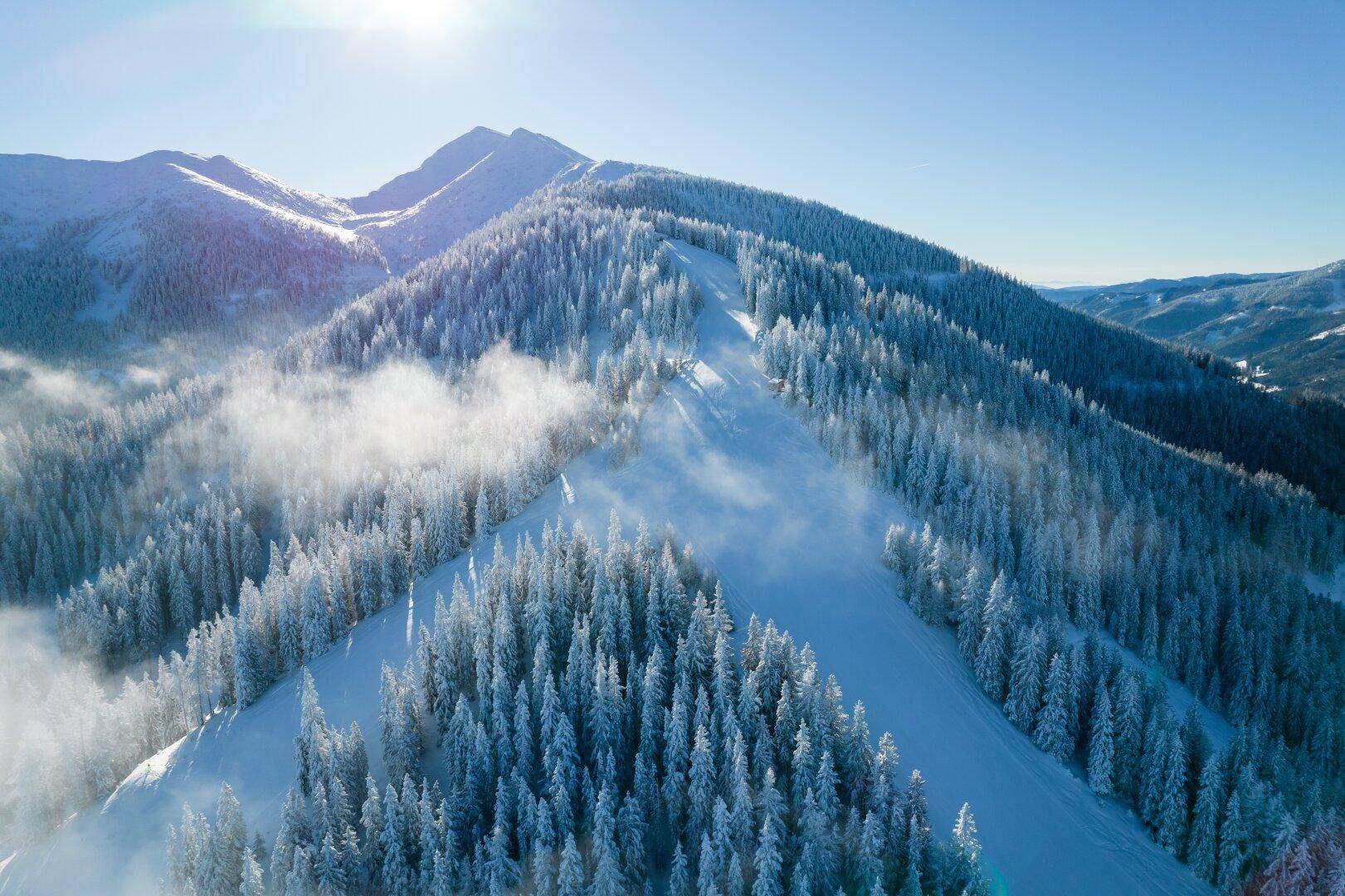 Sonnige Berglandschaft mit verschneiten Wäldern und aufsteigendem Nebel, der die Gipfel umhüllt.