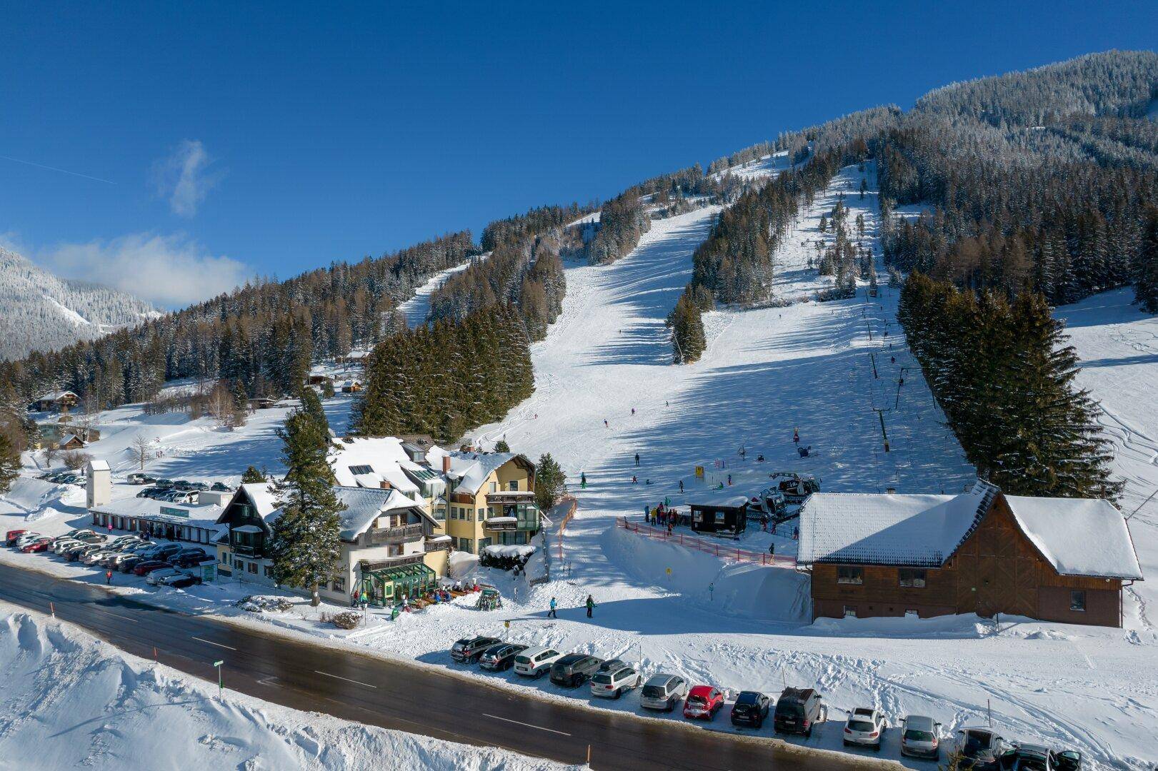 Hotelanlage im Winter mit verschneiten Dächern, umgeben von Skipisten und Parkplätzen.