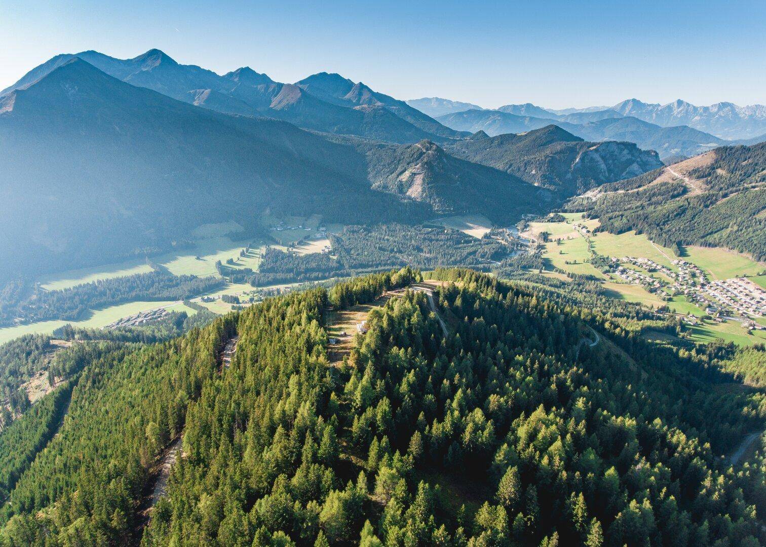 Weitläufige Sommerlandschaft mit grünen Bergen, Wäldern und einem Dorf im Tal.