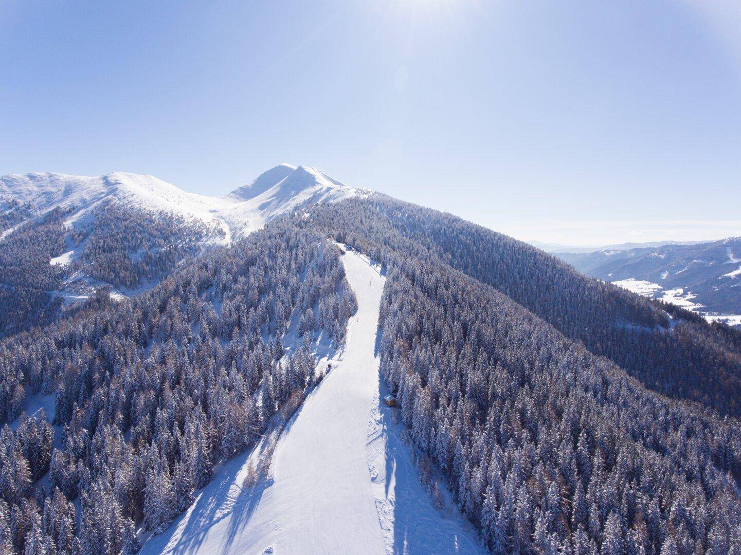 Eine lange, präparierte Skipiste führt durch einen verschneiten Wald mit sonnigem Bergblick.