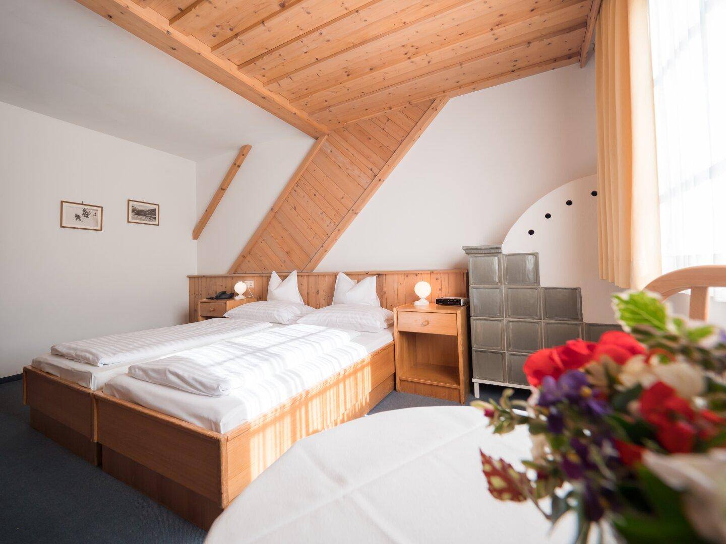 Rustikales Schlafzimmer mit Holzdecke, Kachelofen und einem Blumenstrauß auf dem Tisch.