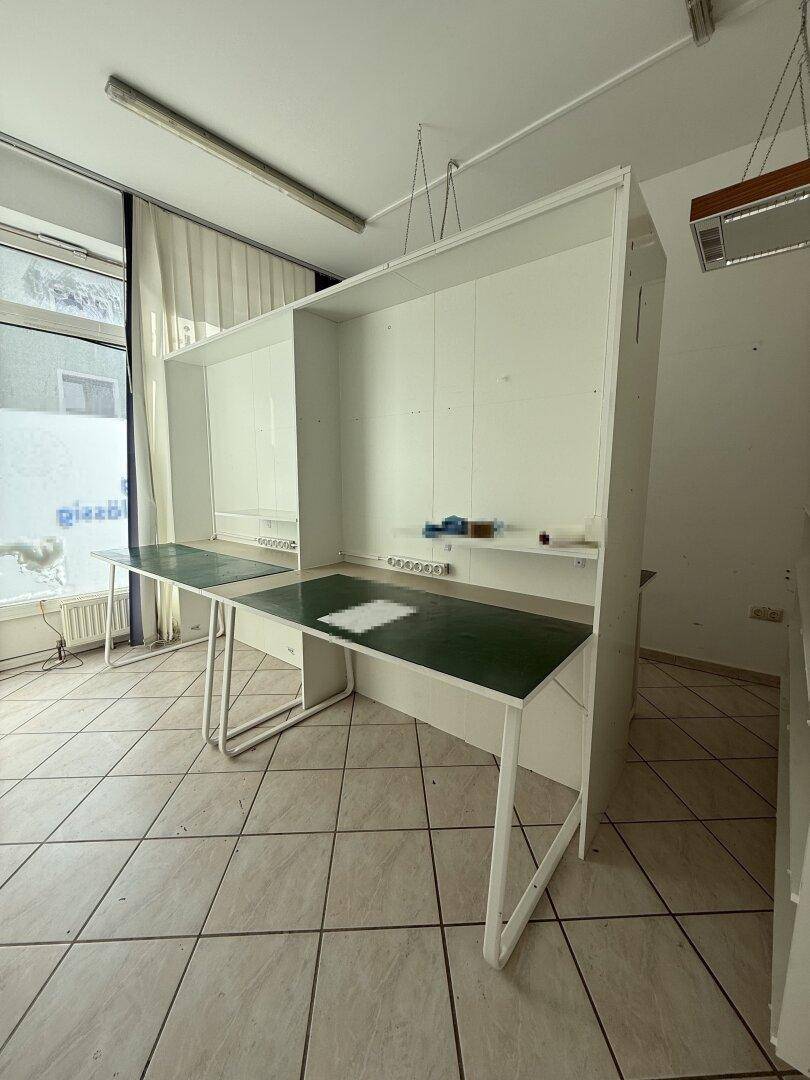 Innenansicht eines Bürobereichs mit Arbeitsplätzen, Fliesenboden und großen Fenstern für viel Tageslicht.