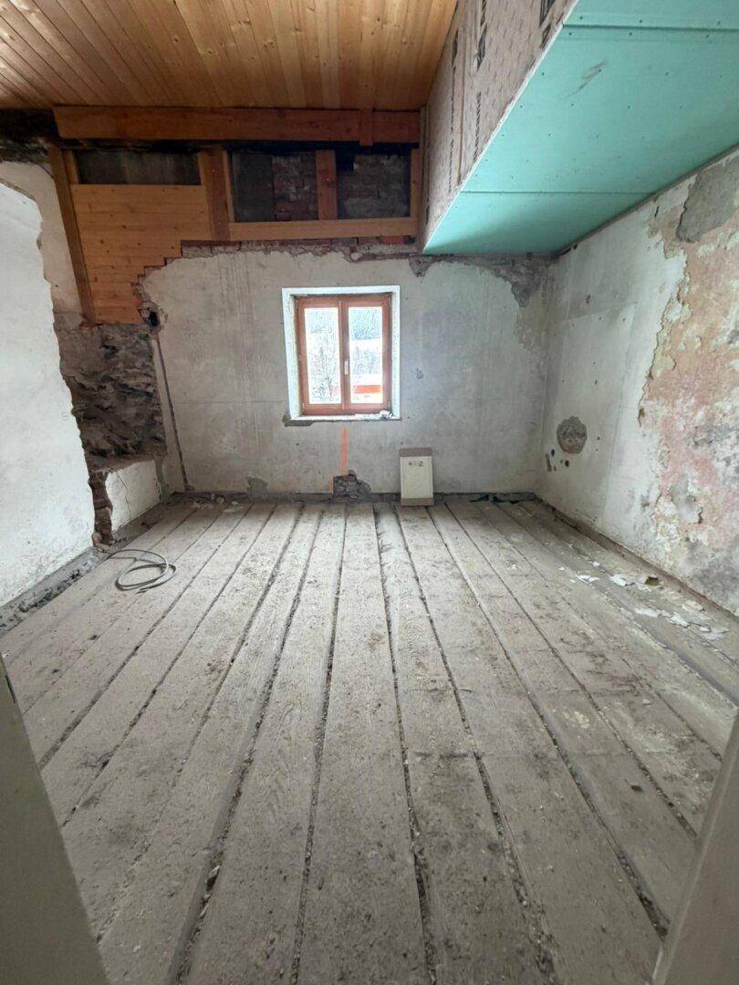 Raum im Rohbau mit alten Holzdielen, einem Fenster und sichtbaren Renovierungsarbeiten.