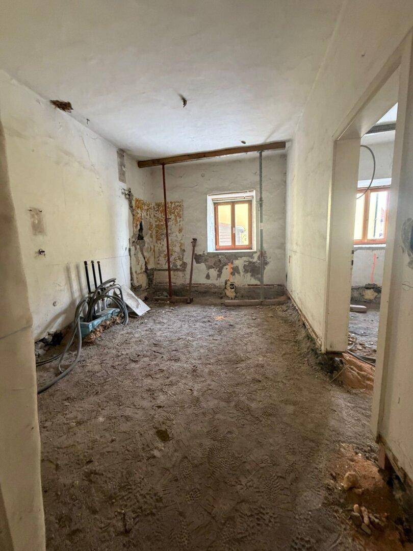 Raum im Rohbau mit sichtbaren Rohrleitungen, einem Fenster und vorbereitetem Boden für den Ausbau.
