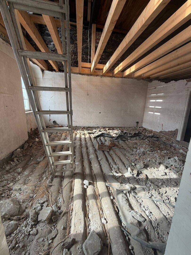 Raum im Rohbau mit freiliegenden Deckenbalken, Schutt auf dem Boden und einer Leiter.