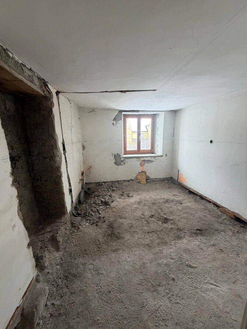Raum im Rohbau mit einem Fenster, sichtbaren Wänden und vorbereitetem Boden für den Ausbau.
