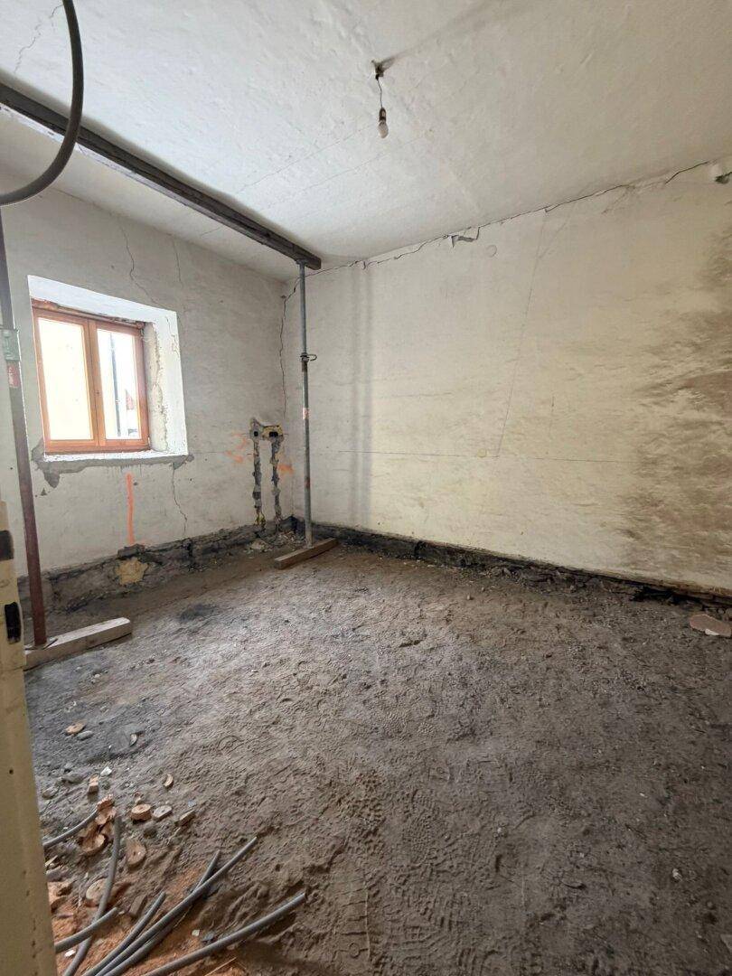 Raum im Rohbau mit einem Fenster, sichtbaren Rohrleitungen und vorbereitetem Boden für den Ausbau.