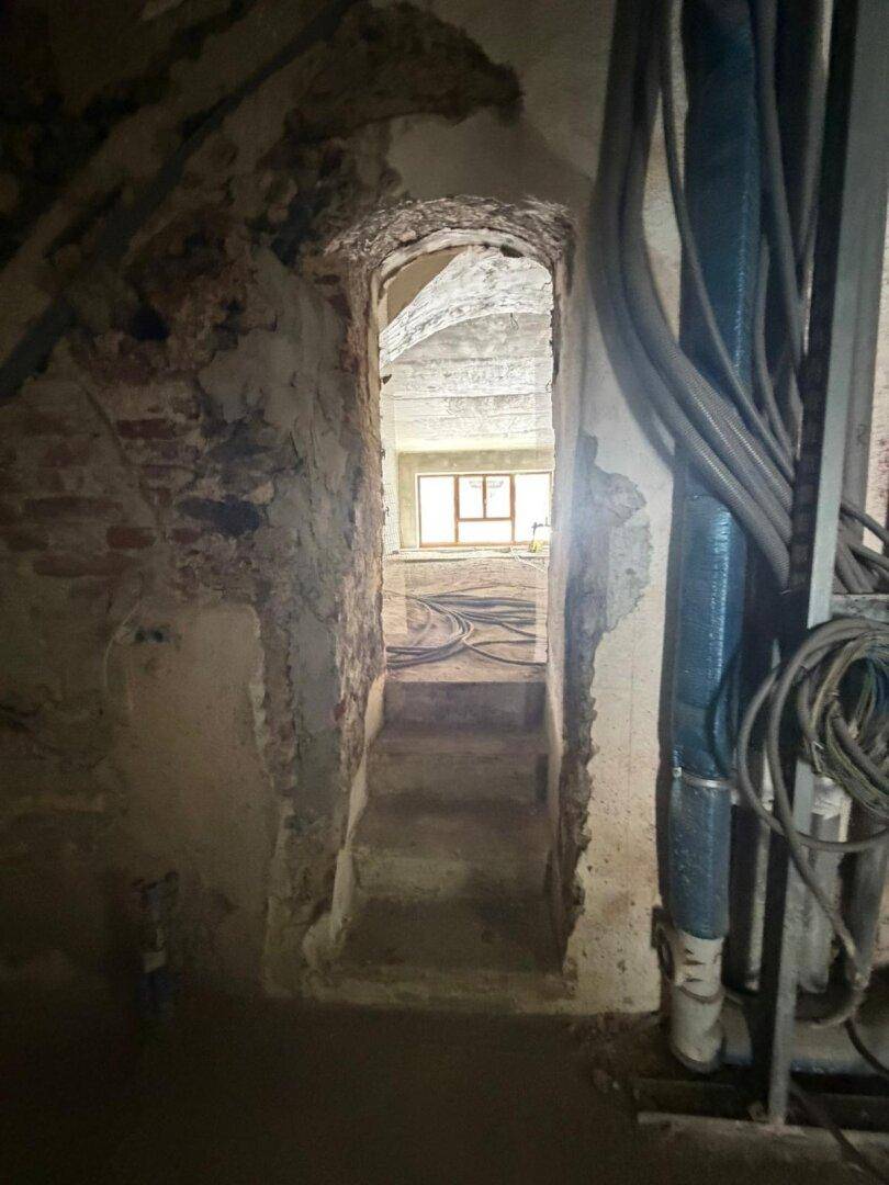 Treppenaufgang im Rohbau mit sichtbaren Installationen und Blick in einen weiteren Raum.