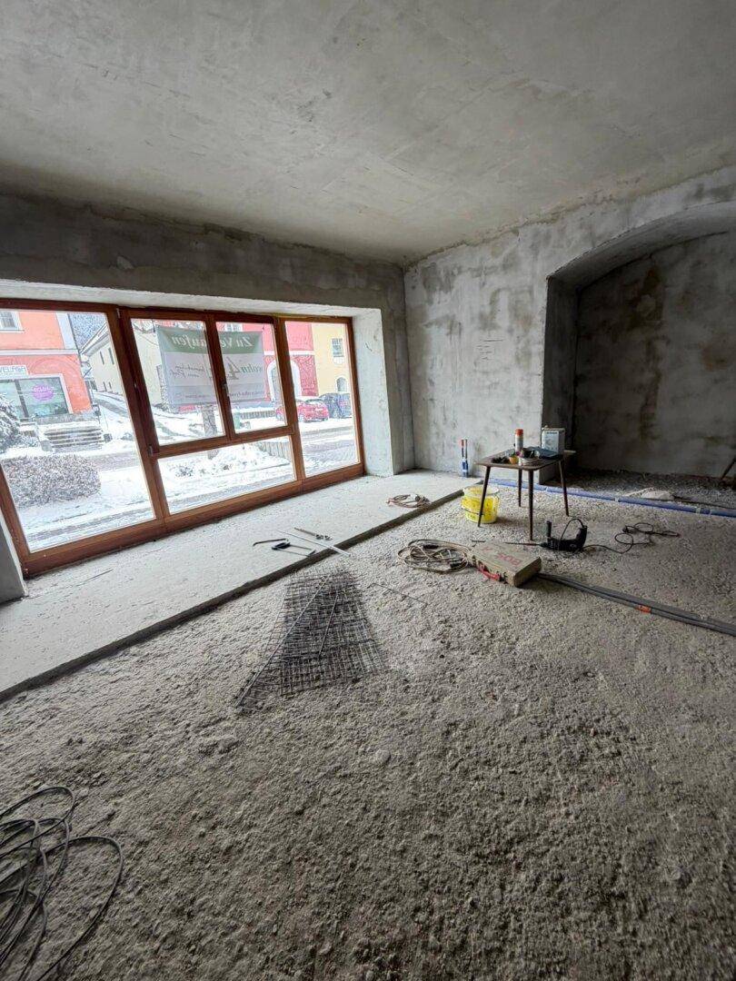 Unfertiger Innenraum mit großen Holzfenstern, Betonboden und Blick auf die Straße, bereit für den Ausbau.