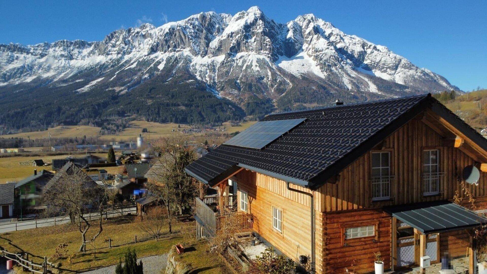 Holzhaus mit Photovoltaikanlage auf dem Dach und Panoramablick auf schneebedeckte Berge und das Tal.