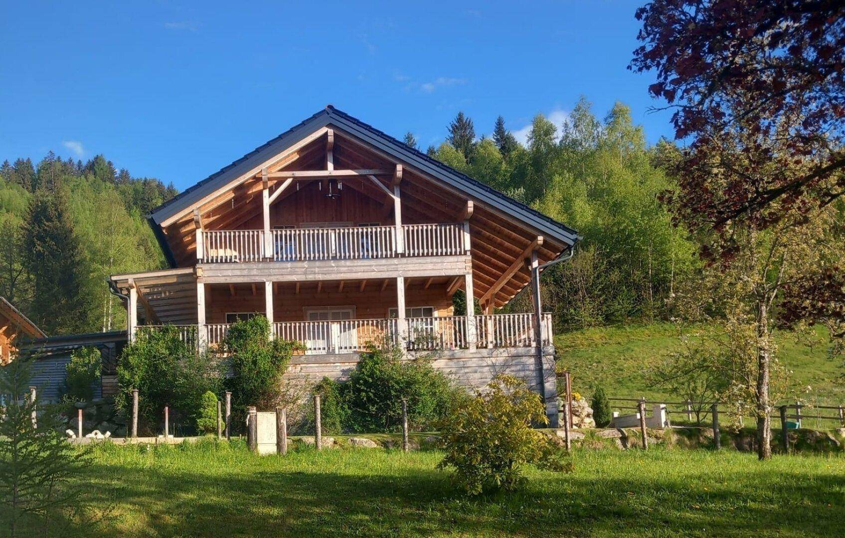 Charmantes Holzhaus mit Balkonen, umgeben von grünen Bäumen und Wiesen an einem sonnigen Tag.