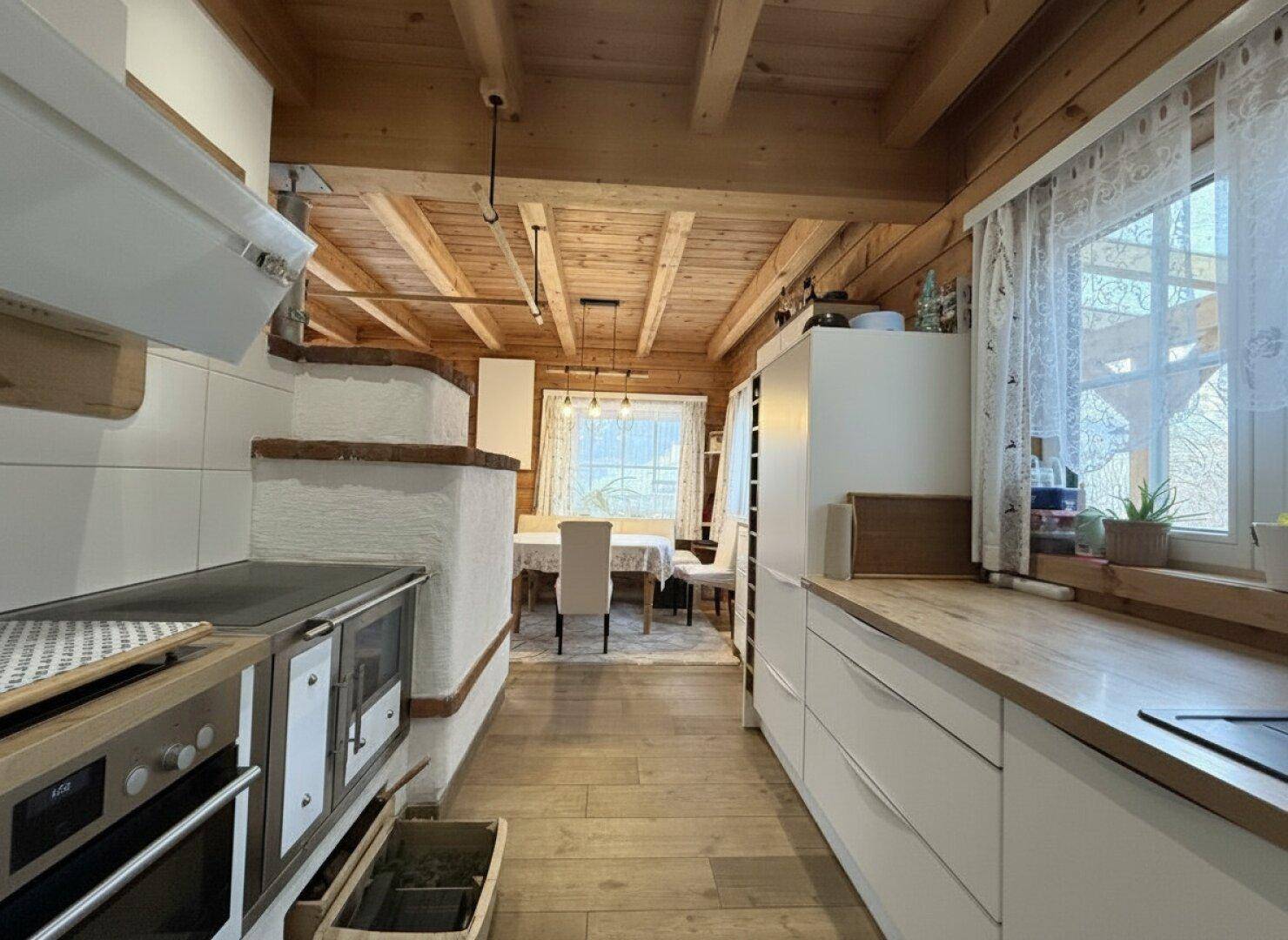 Offene Küche mit Holzbalkendecke und Blick auf den Essbereich mit traditionellem Kachelofen.