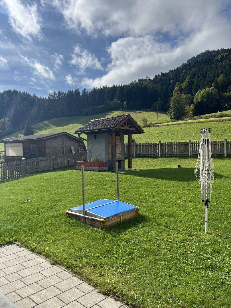 Großer Garten mit Rasenfläche, einem kleinen Holzbrunnenhaus und einem abgedeckten Sandkasten oder Pool.