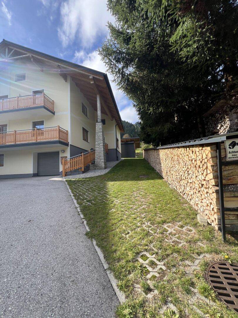 Zufahrt zum Haus mit großem Holzstapel entlang der Mauer und Blick auf die Fassade.