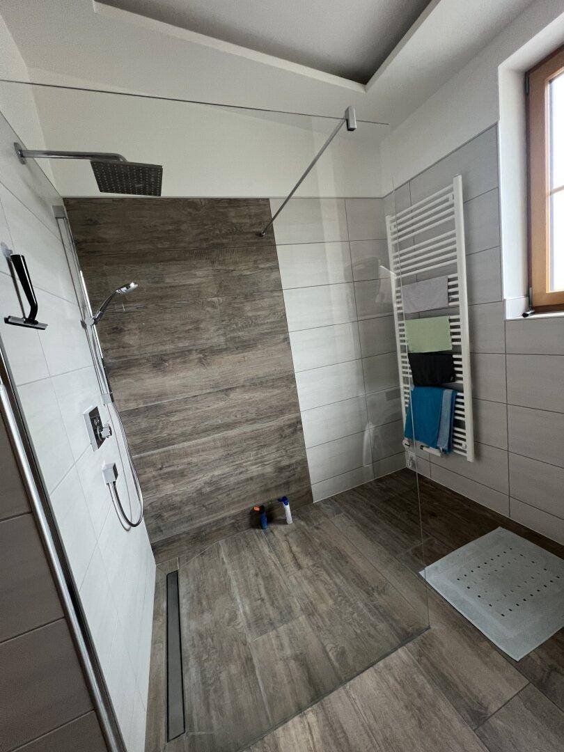 Geräumige, ebenerdige Dusche mit Holzoptik-Fliesen, Regendusche und beheiztem Handtuchhalter.